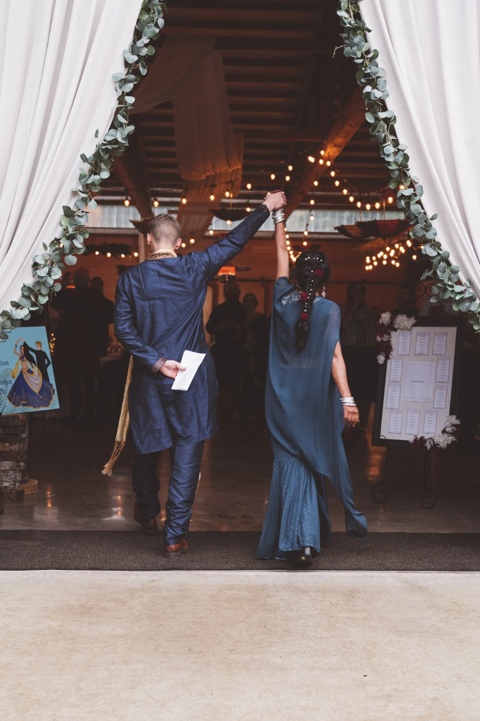 A couple dancing at their wedding reception, holding hands up, with guests in the background and warm string lights overhead.