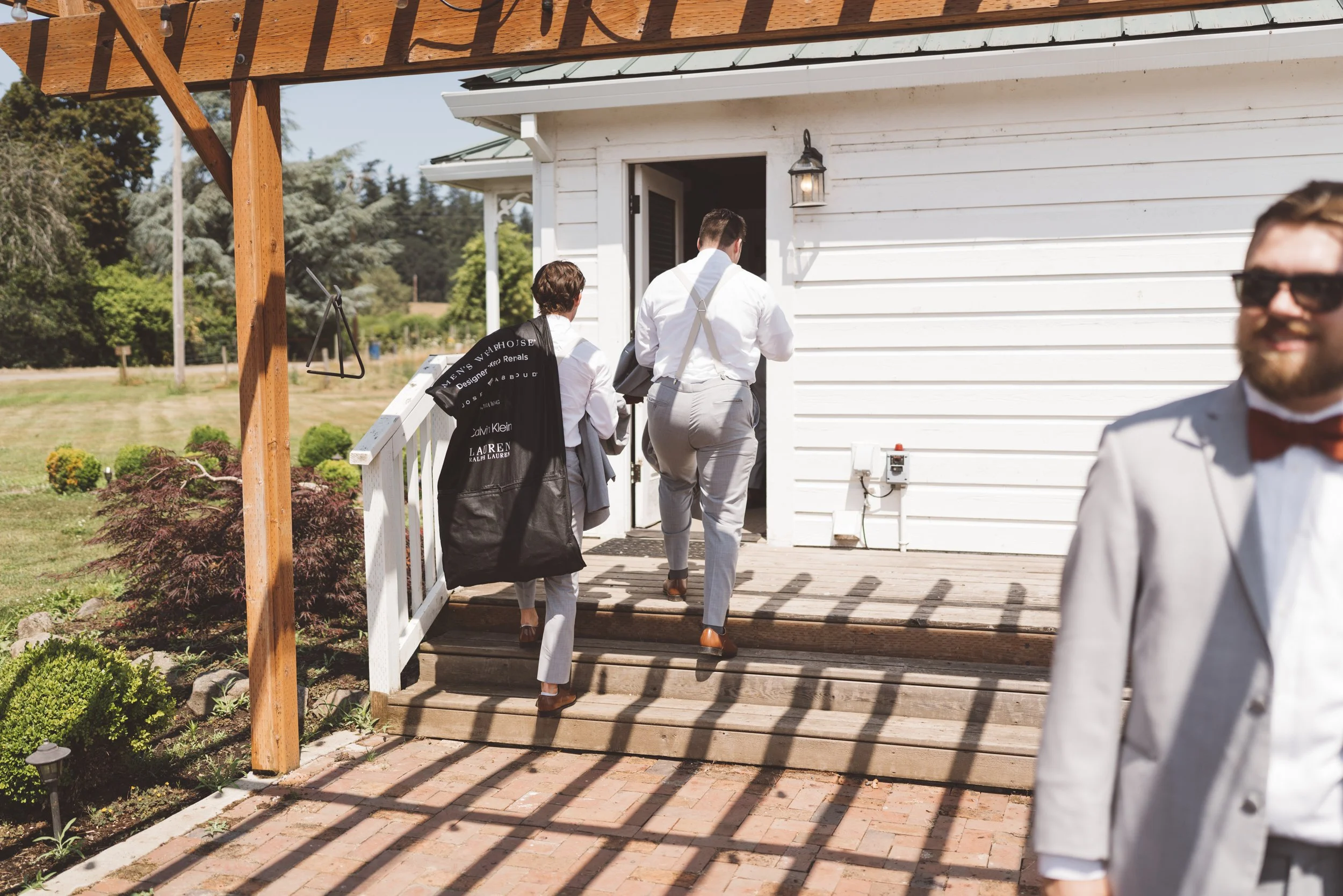 Three men in suits entering a white house on a wooden deck, with a garden and trees in the background, during daytime.