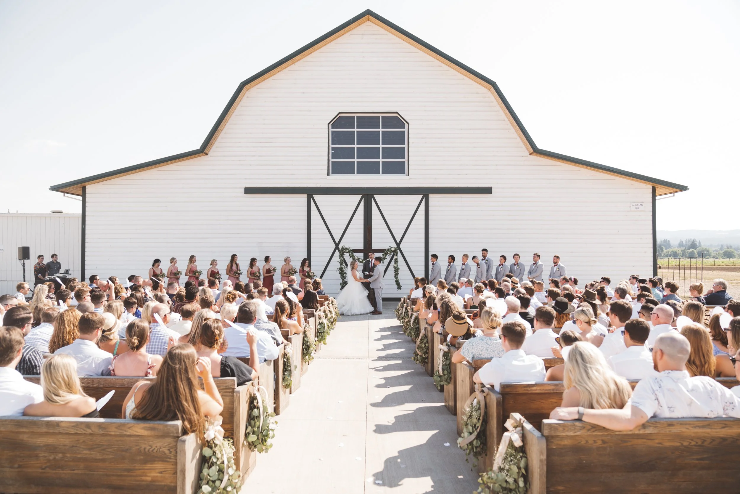 Outdoor wedding ceremony with a bride and groom exchanging vows in front of a large white barn, surrounded by friends and family seated on wooden benches, with bridesmaids and groomsmen on either side.