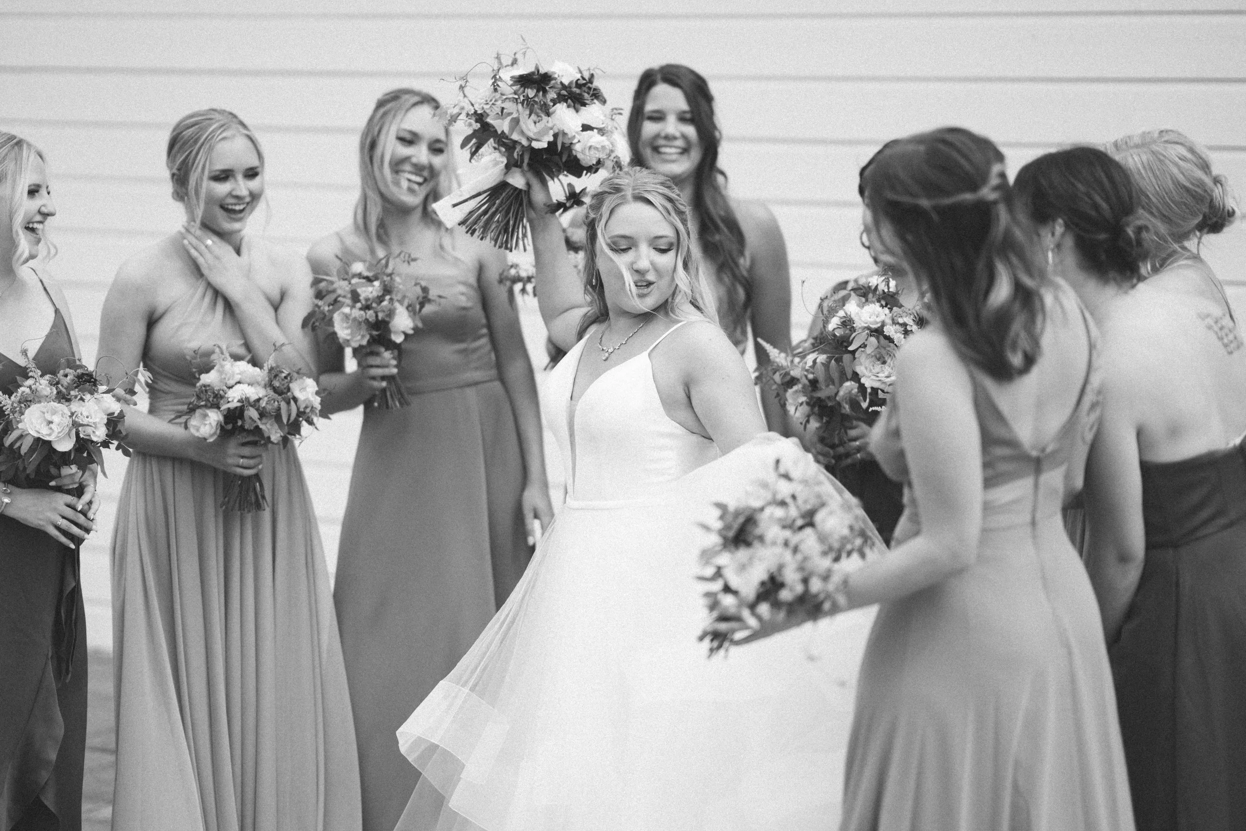 A bride in a white wedding dress surrounded by bridesmaids holding bouquets, at a wedding celebration.