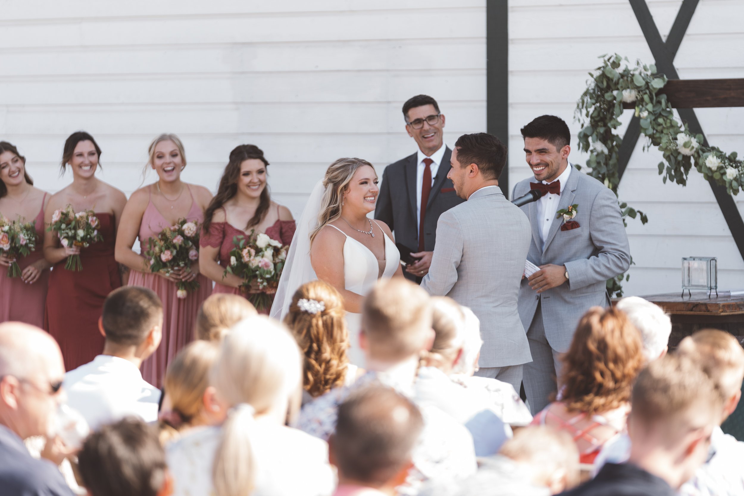 A wedding ceremony with a bride and groom exchanging vows, surrounded by bridesmaids and guests, outdoors in front of a white wooden wall decorated with greenery.
