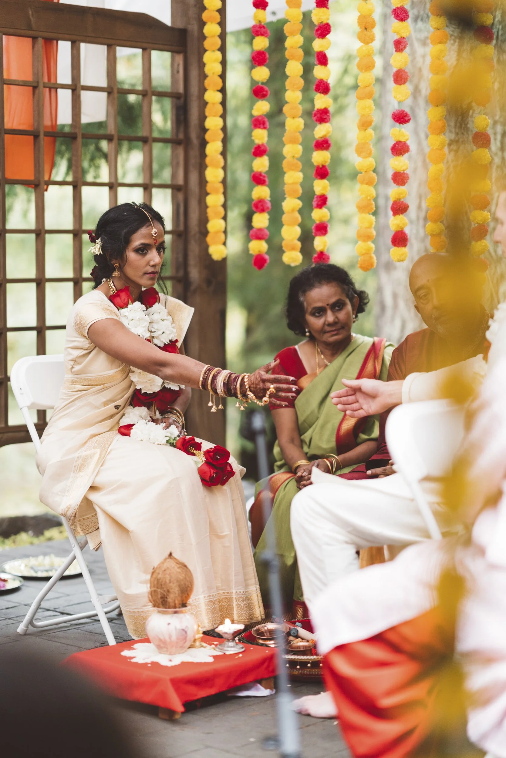 A woman dressed in traditional Indian attire, sitting outdoors during a ceremony, reaching out her hand towards an unseen person, with colorful flower garlands hanging overhead.