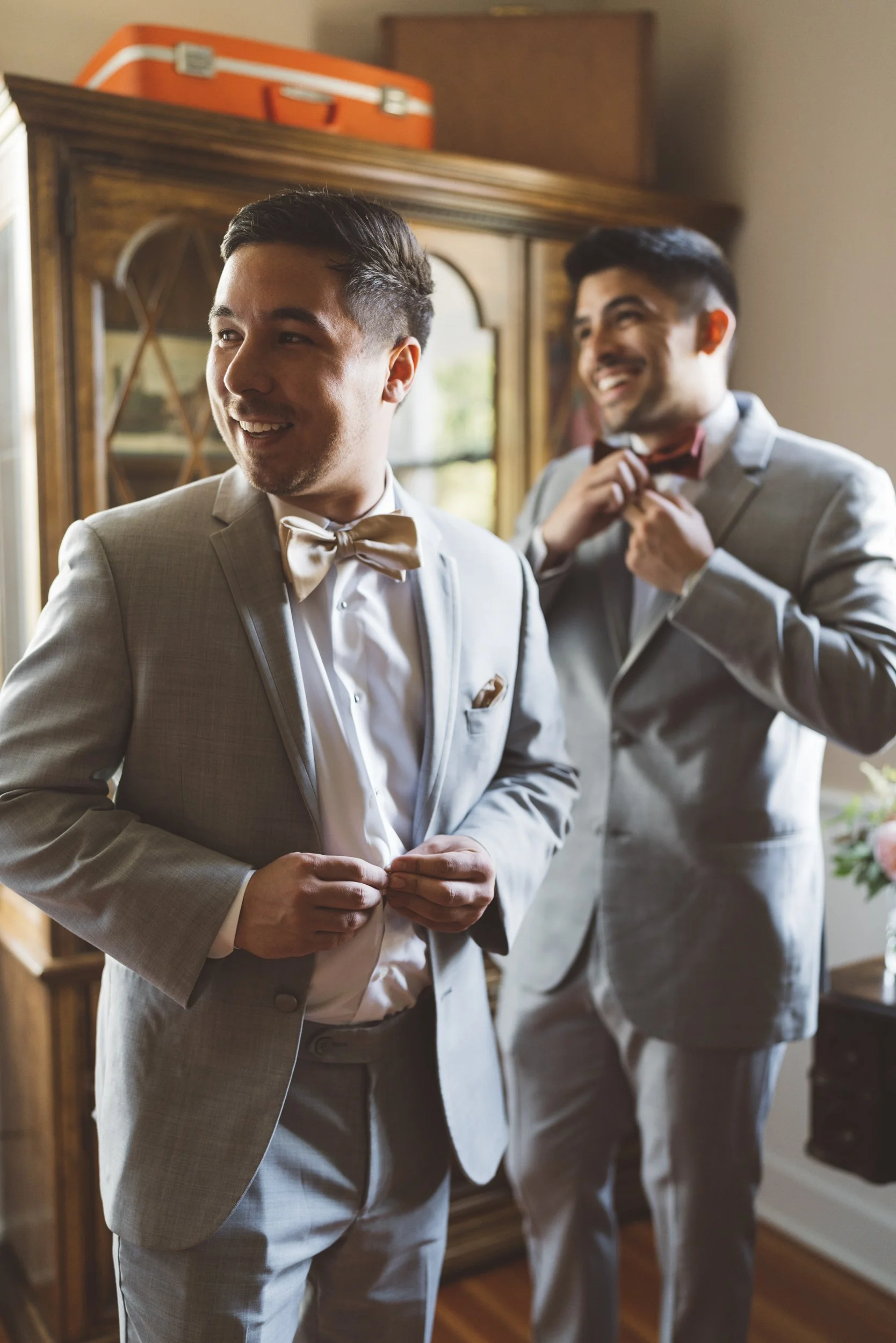 Two men in suits preparing for a formal event, with one adjusting his bowtie and the other buttoning his jacket in a home setting.