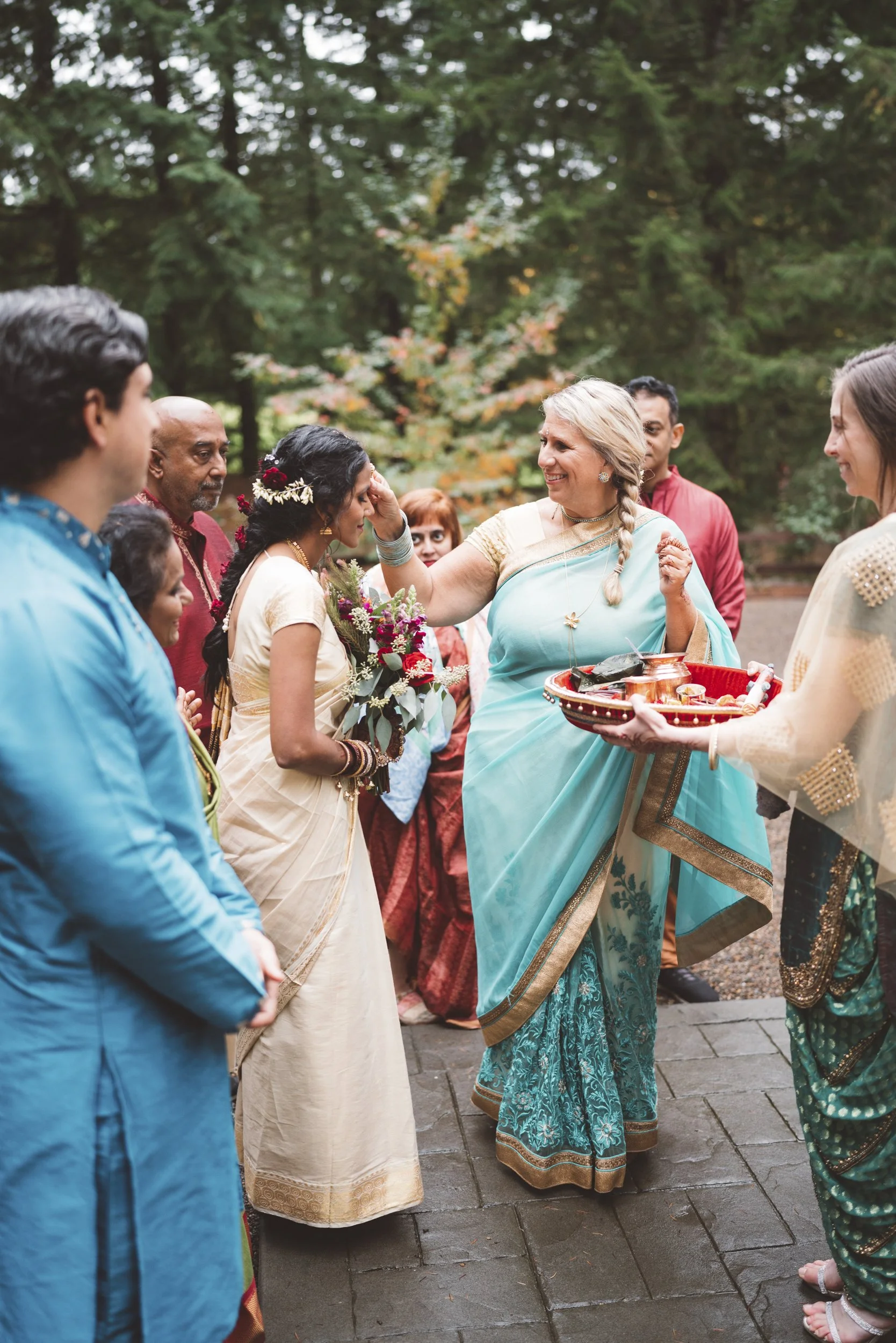 A group of people at an outdoor Indian wedding ceremony. A woman in a blue saree is applying a tilak or mark to the forehead of a woman in a cream saree. Other guests in colorful traditional attire are gathered around, some holding flowers and a tray