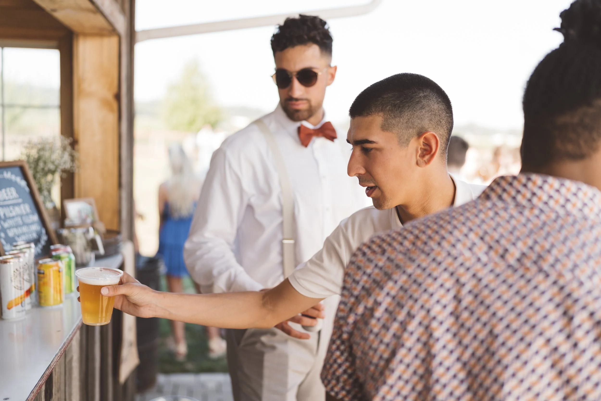 Young man with short hair in a white shirt handing a glass of beer to another person at an outdoor bar with people in the background.
