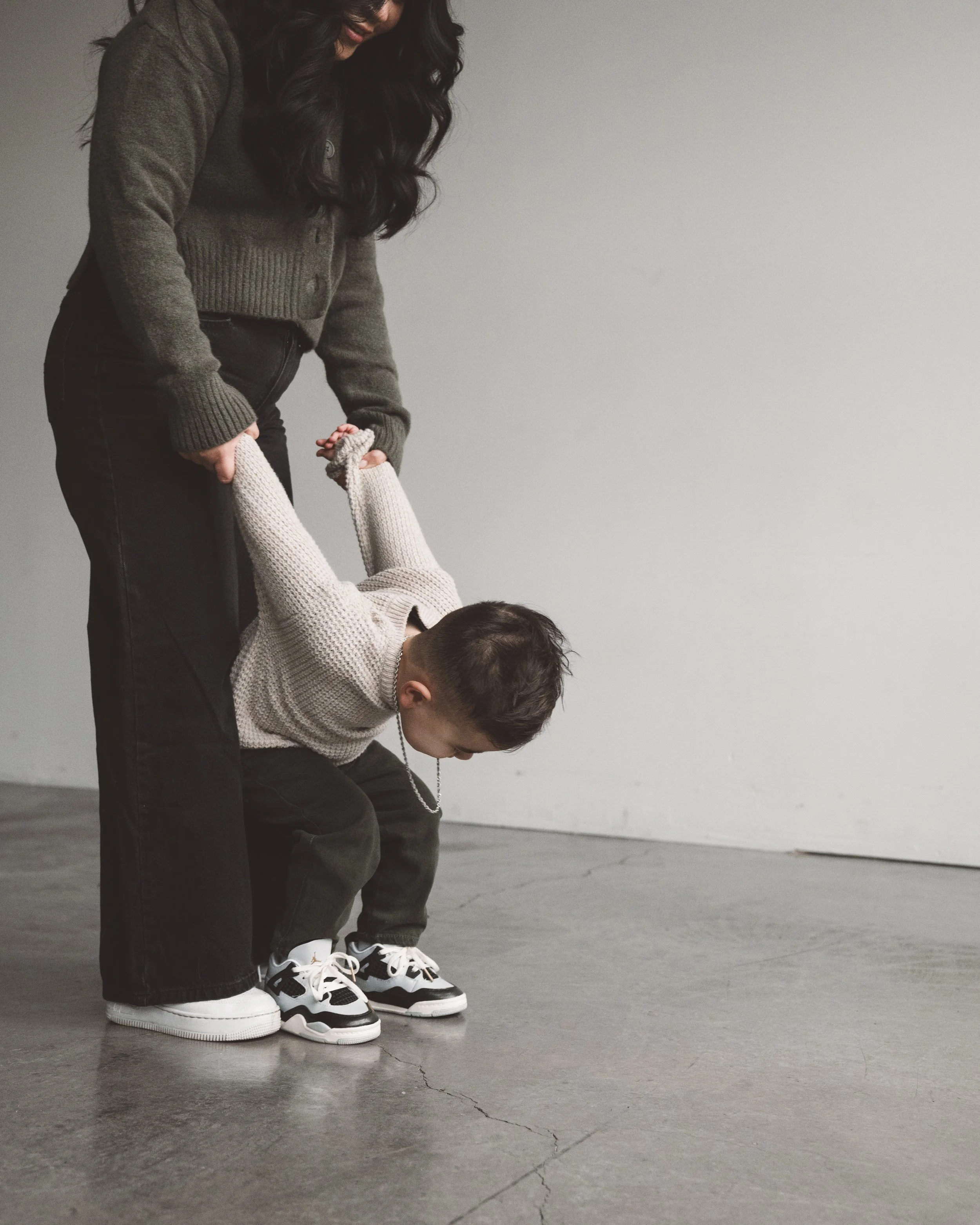A woman holding a young boy's hands as he bends forward, standing on a polished concrete floor against a plain wall.