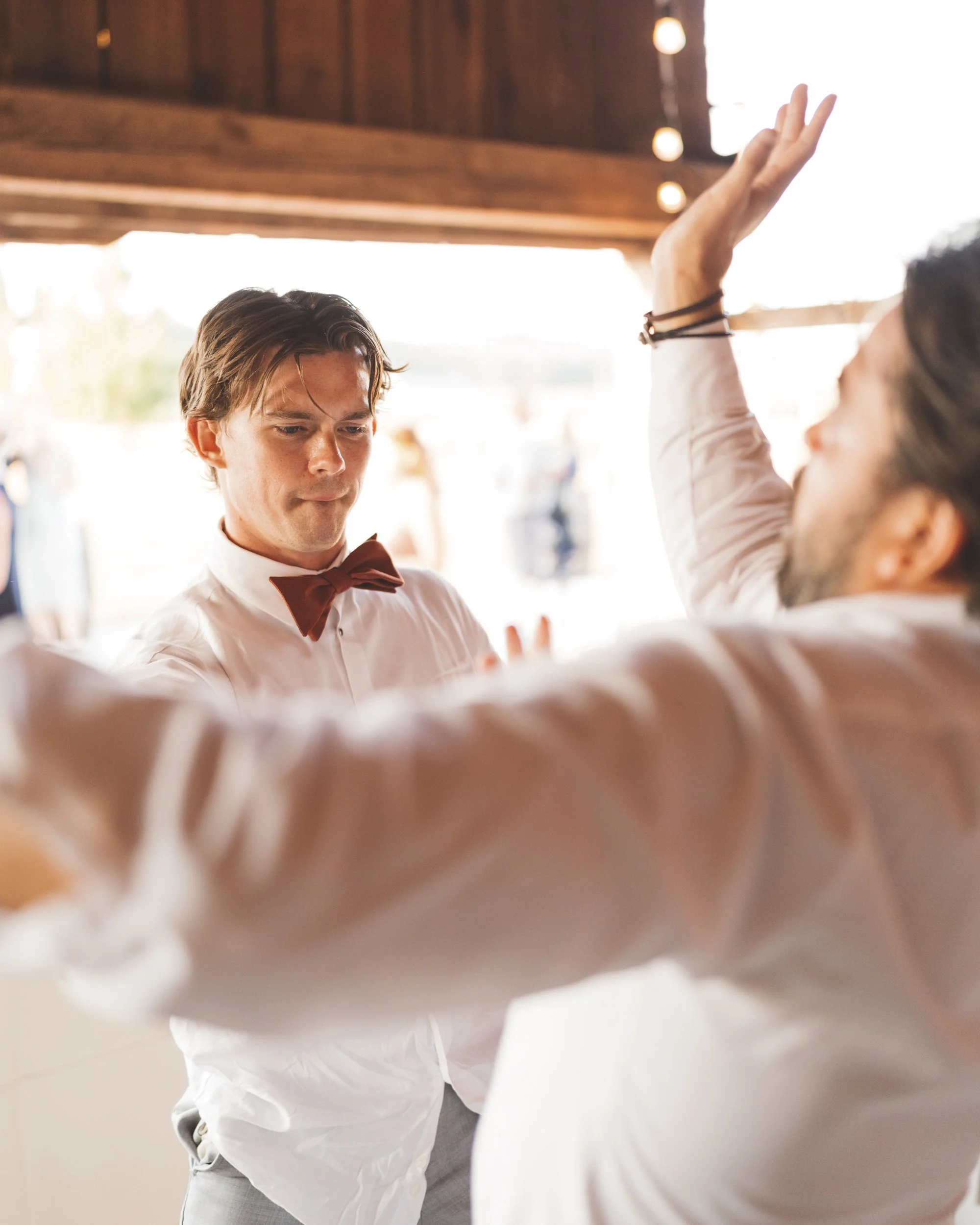 Two young men dancing at an indoor event, wearing formal attire with bow ties. One has his hand raised and the other is looking at him.
