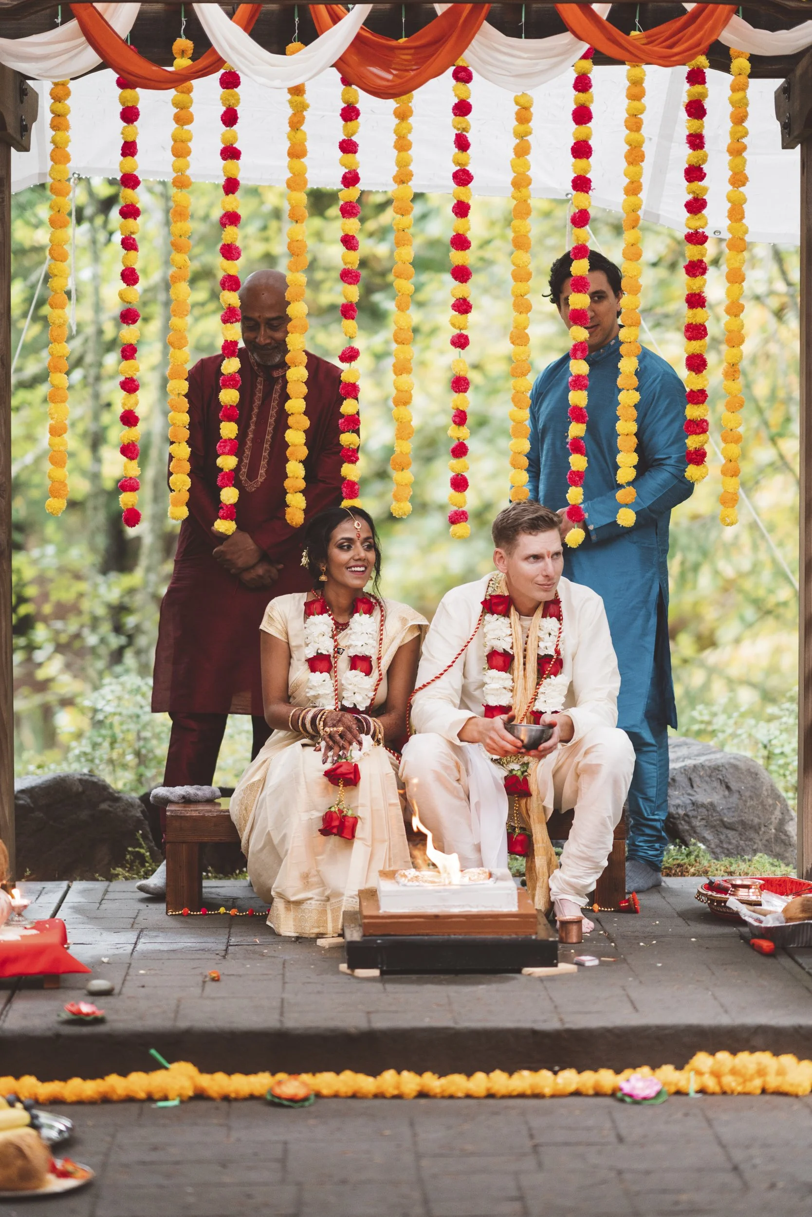 Indian wedding ceremony with bride and groom seated on a stage, the bride in a cream-colored sari and the groom in white, both wearing floral garlands. An officiant and a man in traditional attire stand behind them, with colorful floral decorations h