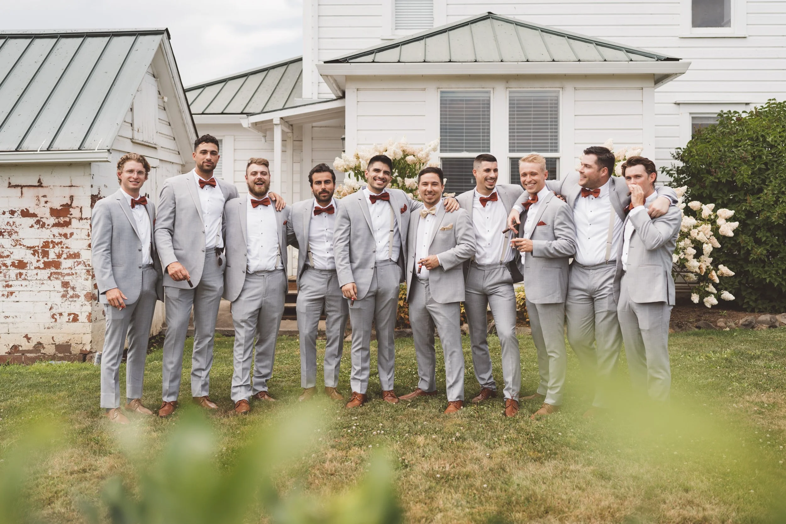 A group of eleven men dressed in light gray suits with brown shoes and red or gold bow ties, standing outside on grass in front of a white house with flowering bushes. They are smiling and posing for a photo, some with arms around each other.