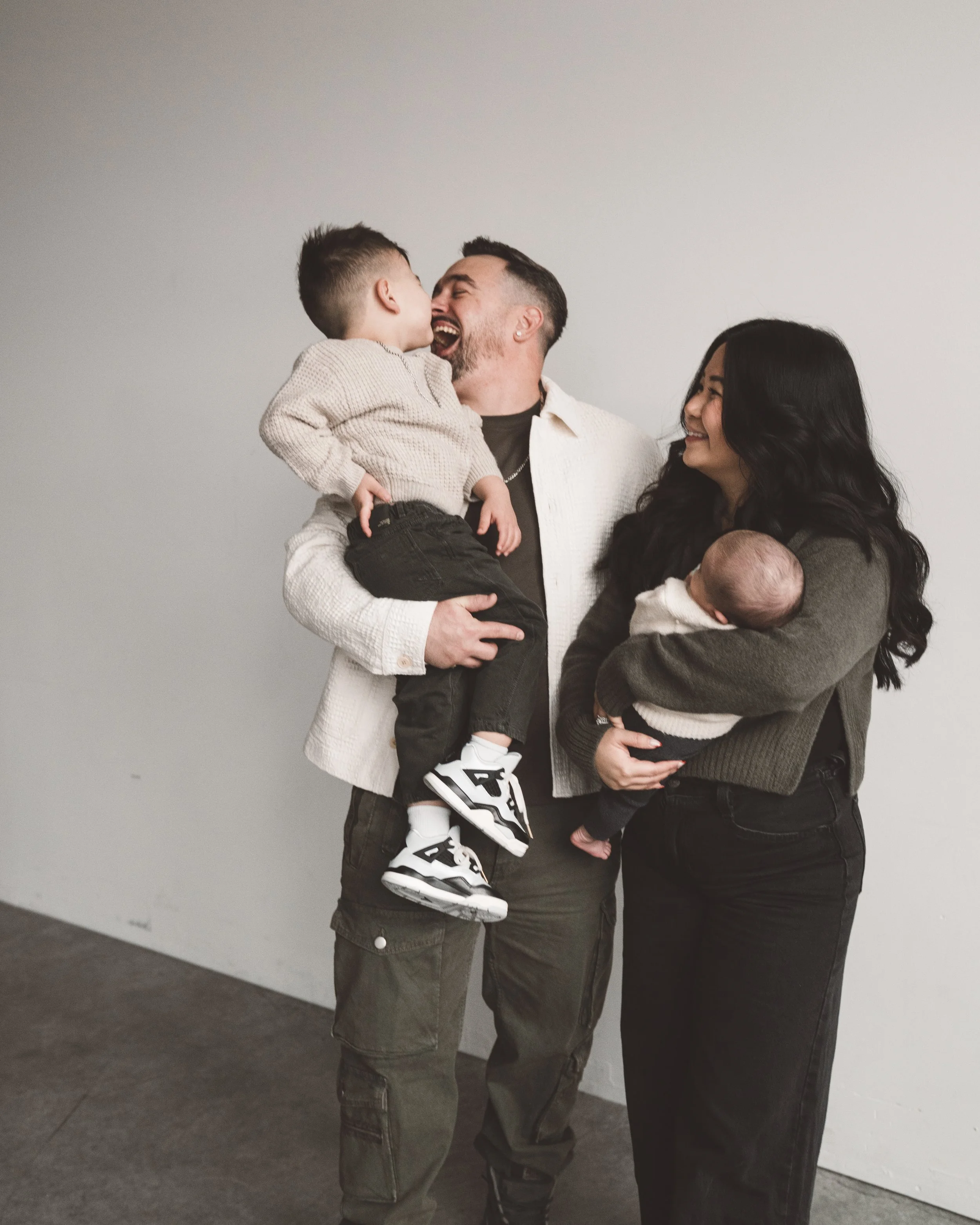 A happy family of four, including a dad, mom, a young boy, and a baby, standing against a plain gray wall in an indoor setting. The dad is holding the boy, who is leaning in to kiss him, while the mom holds the baby and smiles at her family.