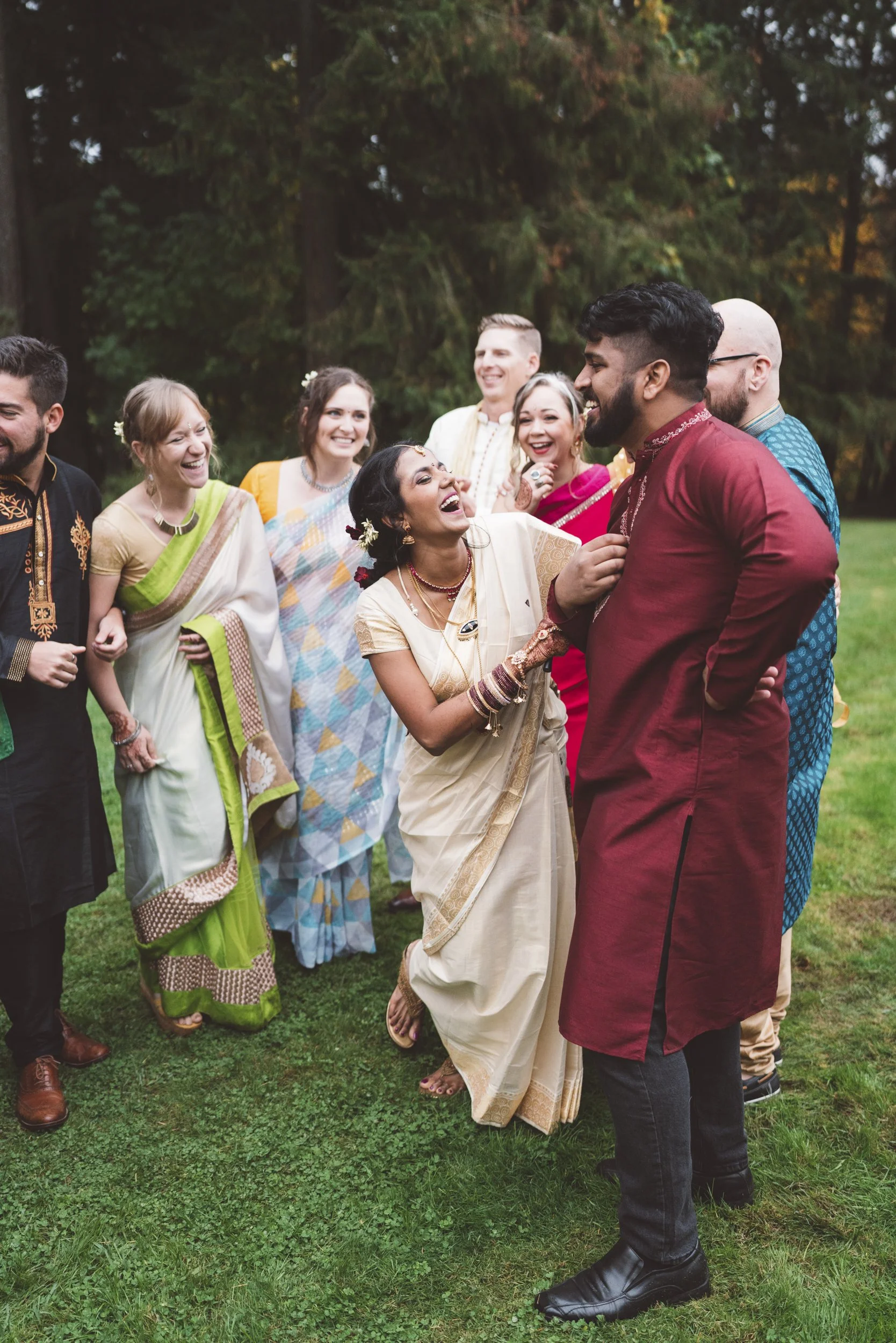A group of people in traditional Indian attire smiling and laughing outdoors on a grassy area with trees in the background.