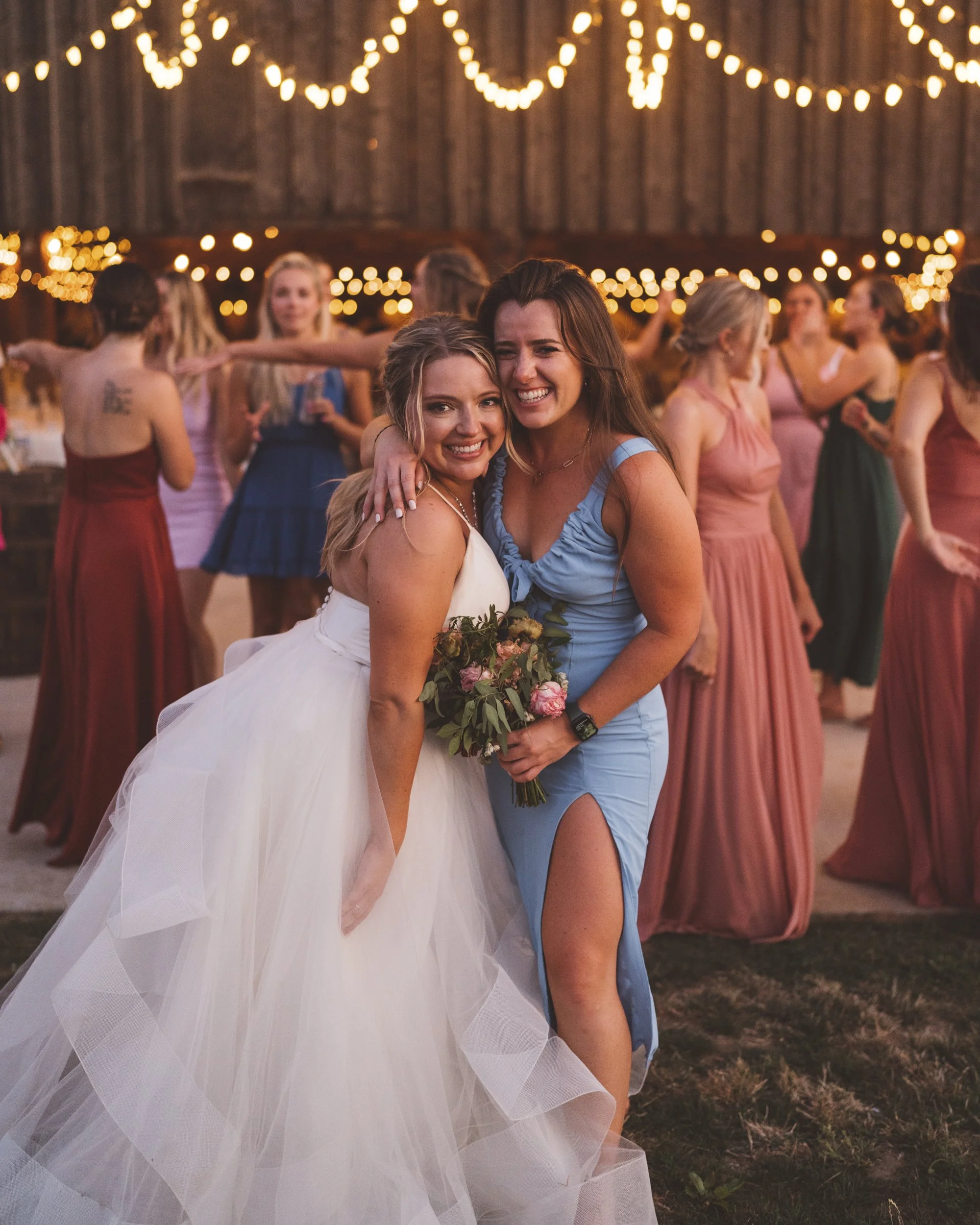 Two women are smiling and hugging at a wedding reception under string lights, with other women in dresses in the background.