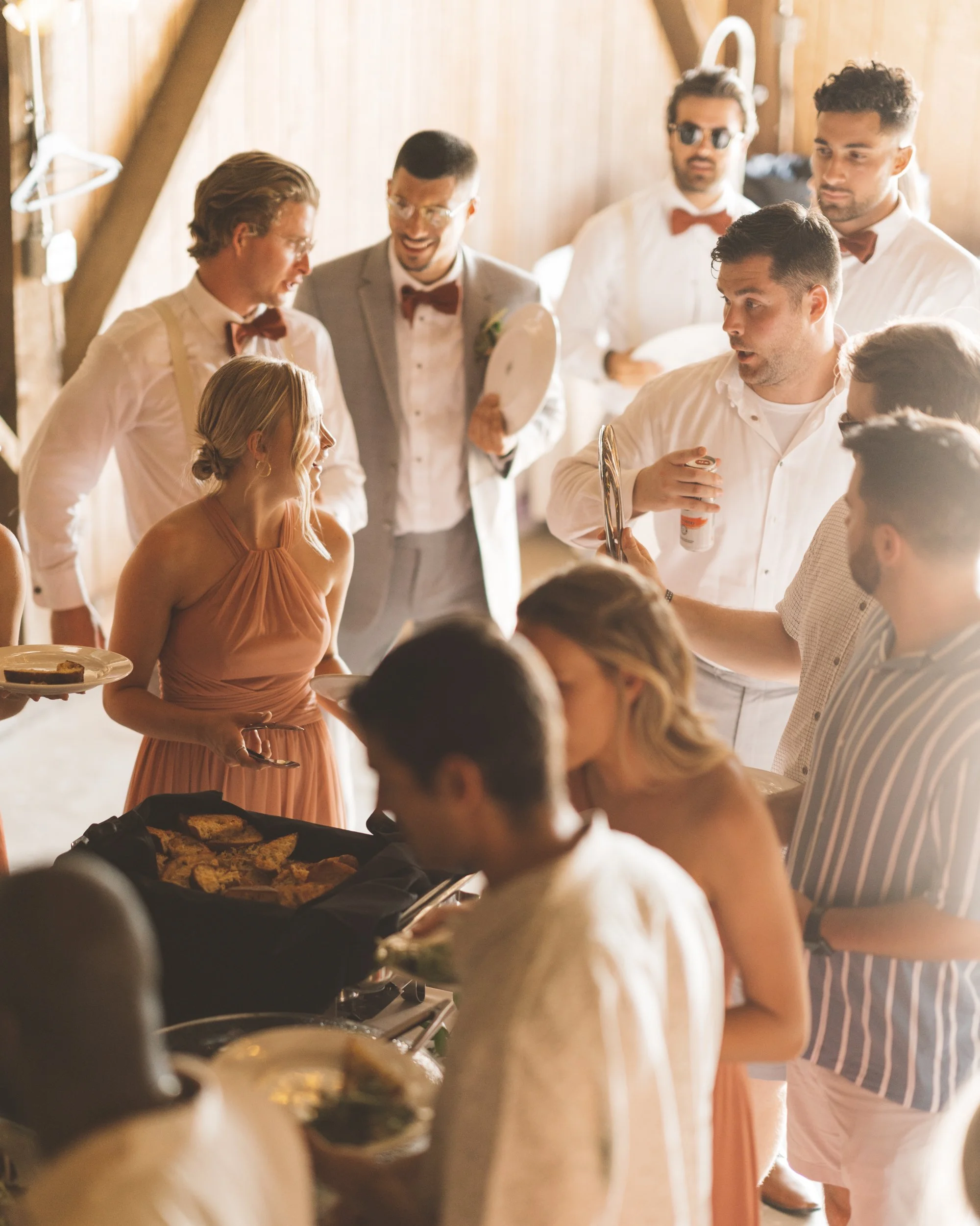 Guests at a casual event serving themselves food and drinks, with a group of men in white shirts, some with bow ties, smiling and talking, a woman in a peach dress holding a plate, in a wooden, rustic setting.