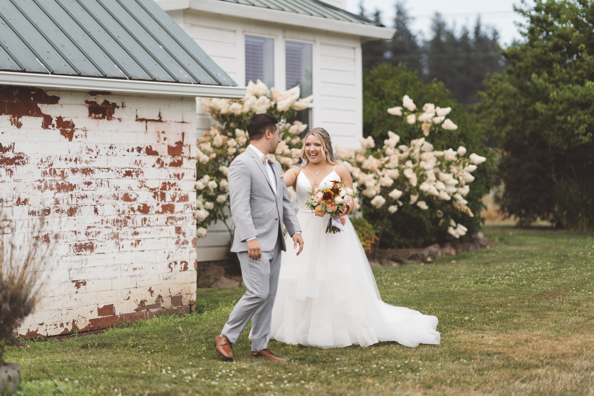 A bride in a white wedding gown holding a bouquet of flowers, standing in front of a groom in a light gray suit outside near a white house with blooming bushes.