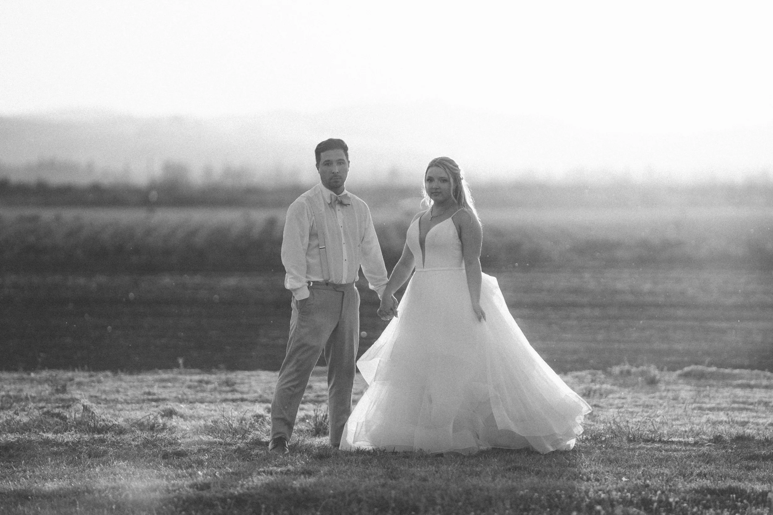 Black-and-white photo of a bride and groom holding hands outdoors in a field, with a blurred landscape in the background.