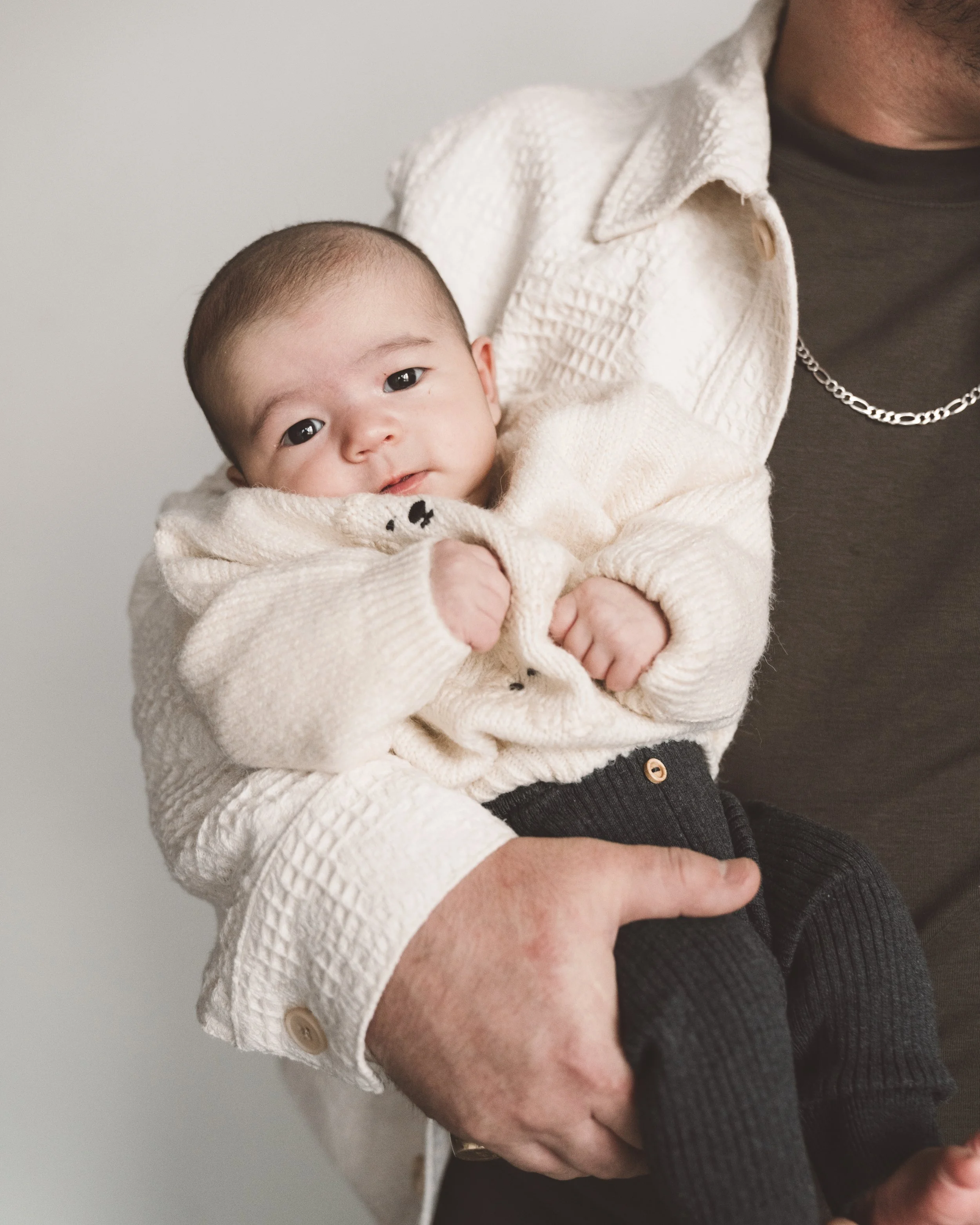 A baby being held by an adult, dressed in a cream sweater and dark pants, looking at the camera with a neutral expression.
