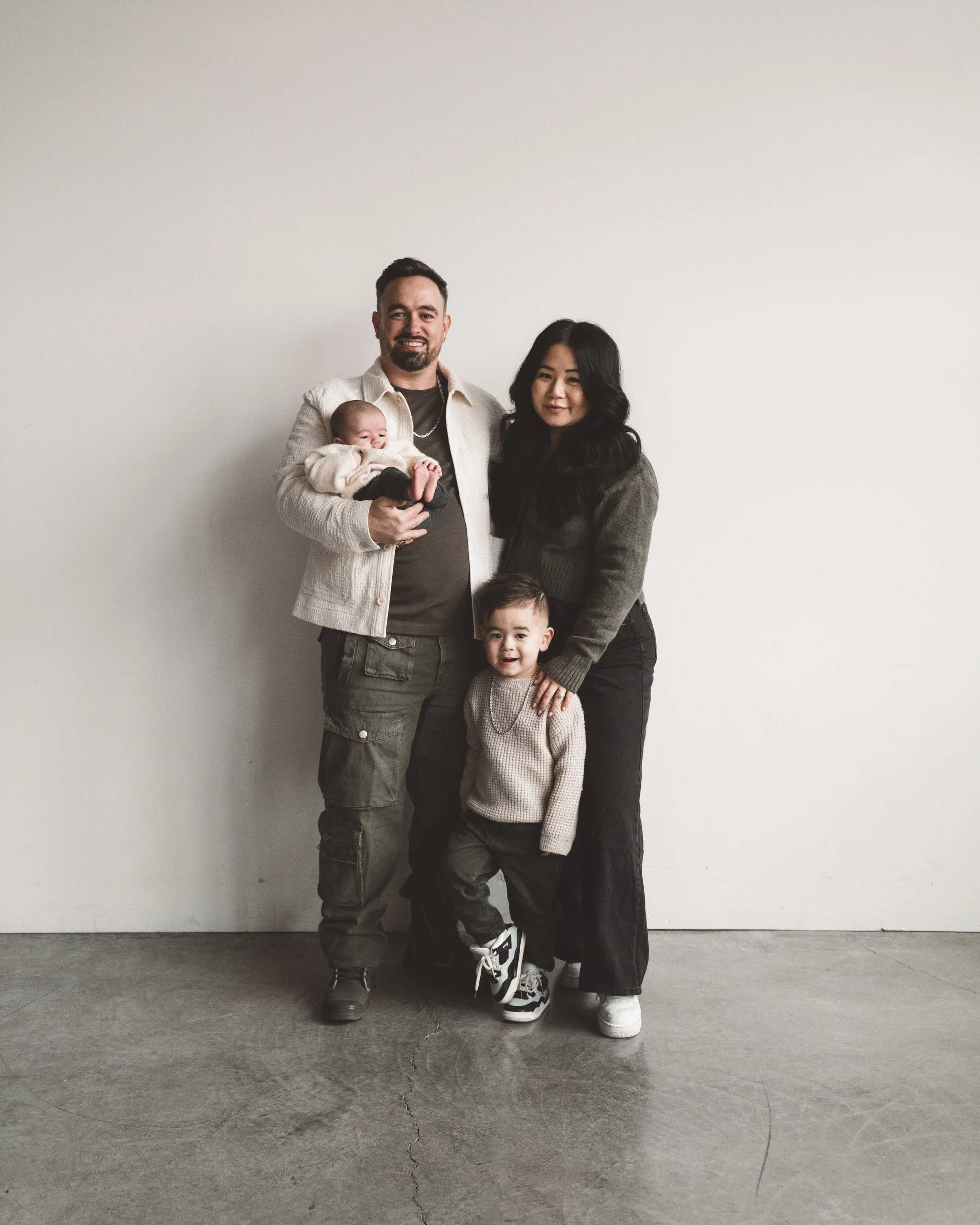A family of four standing together in a room with a white wall background. The father is holding a baby, the mother has her hand on a young child's shoulder, all smiling.