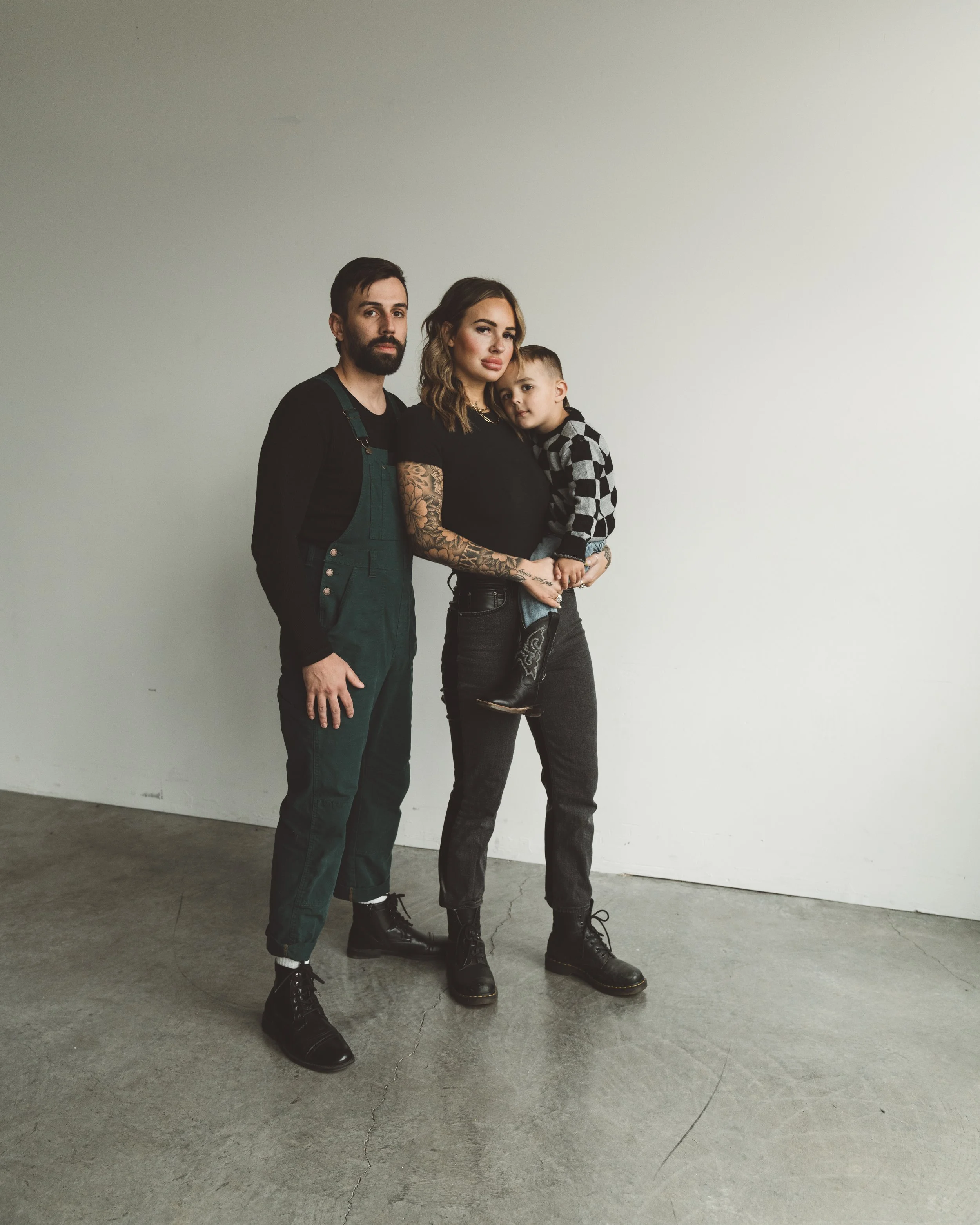 Three people standing together against a plain wall, including a man, a woman, and a young boy, all dressed in casual dark clothing.