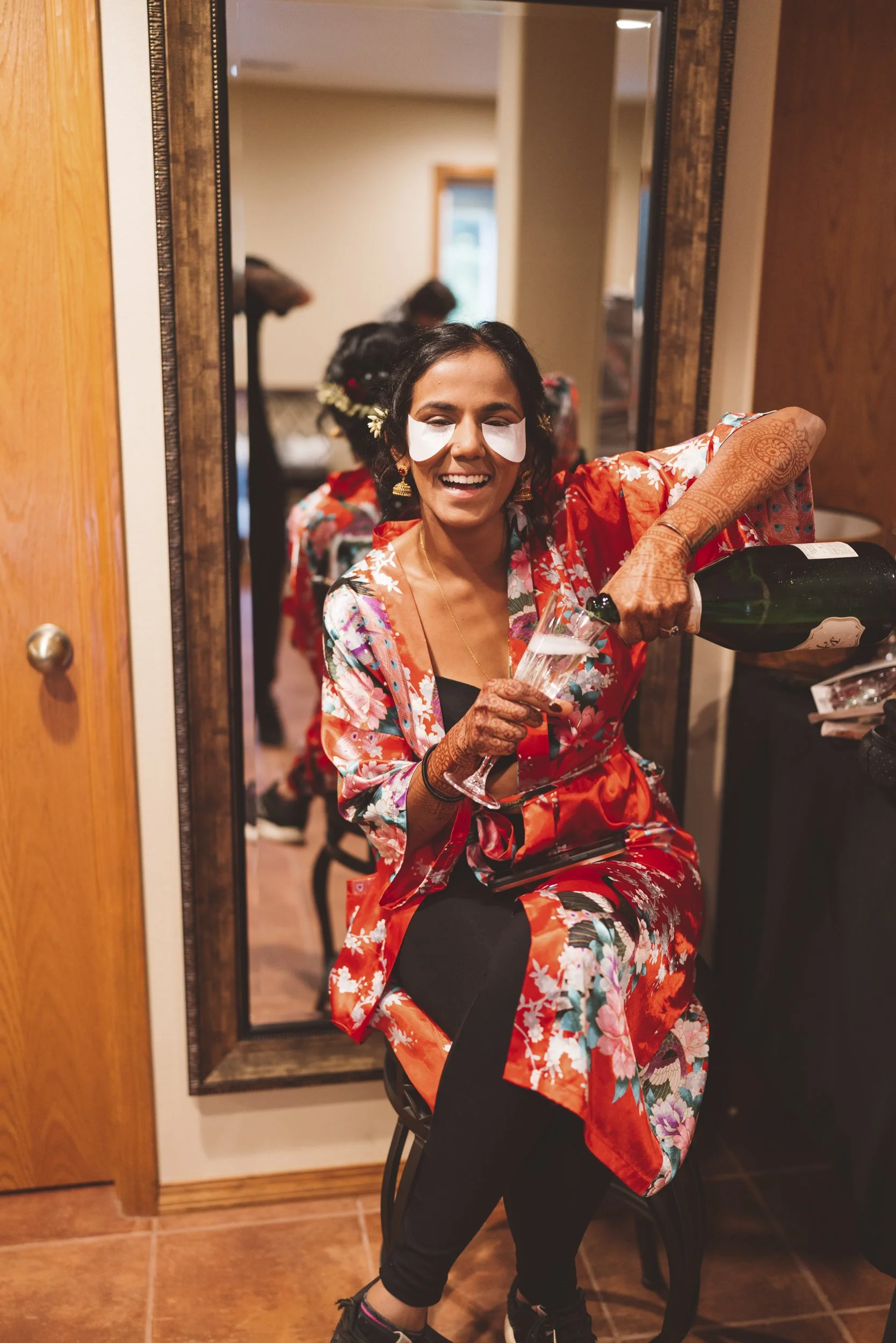Woman in floral kimono pouring champagne into a flute in front of a mirror, celebrating at a gathering.