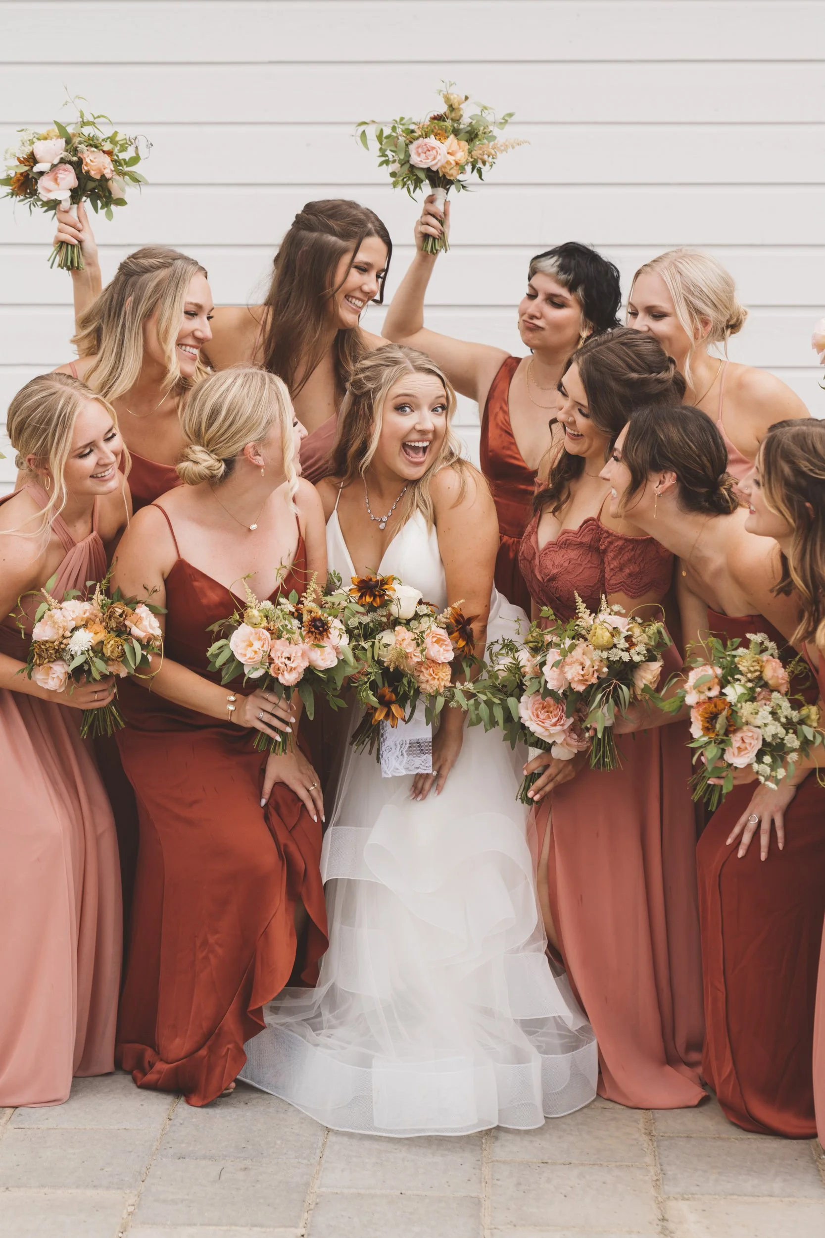A group of women, including a bride in a white dress, surrounded by bridesmaids in shades of pink and red, holding bouquets, smiling and celebrating.