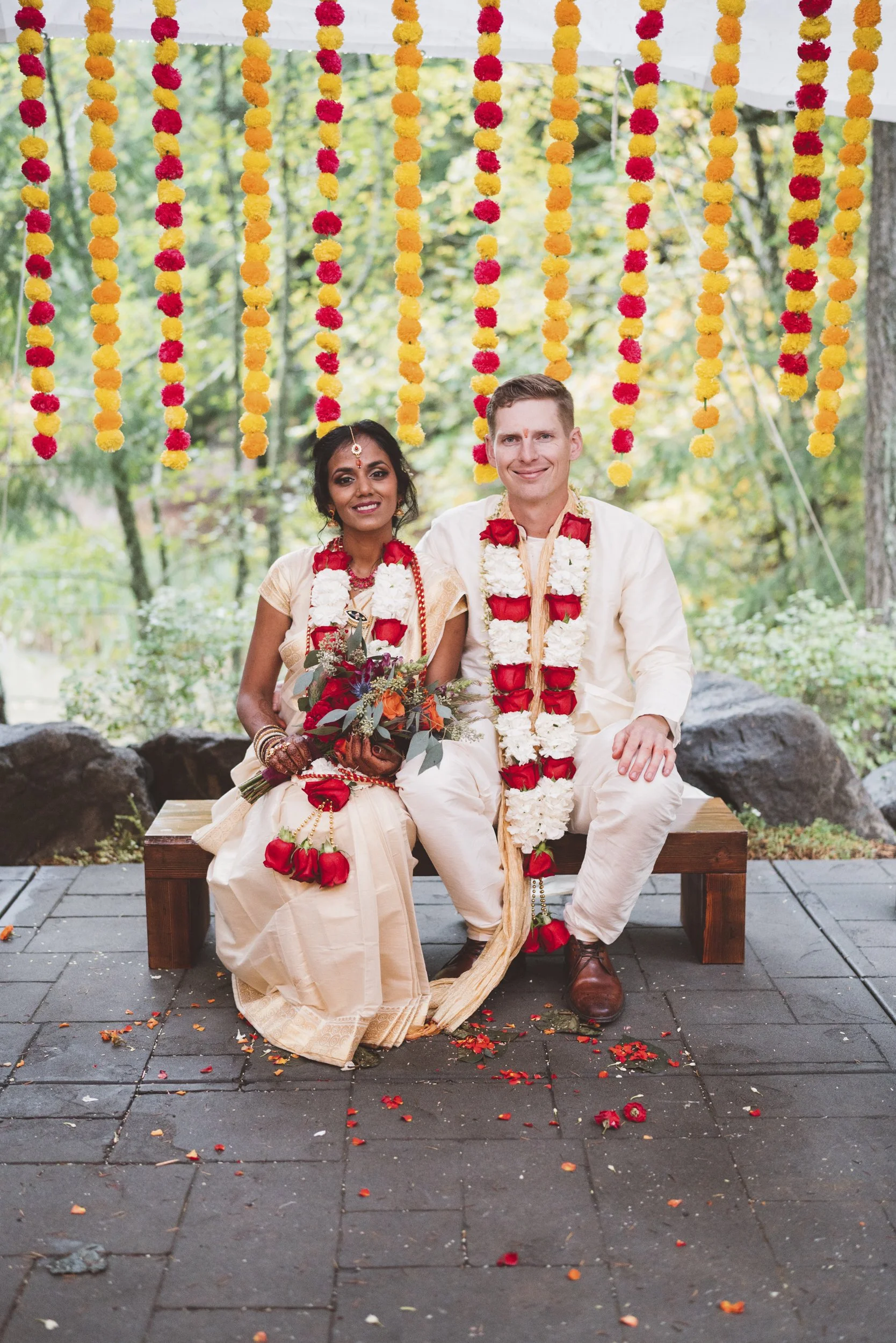 Couple dressed in traditional Indian wedding attire sitting on a wooden bench with floral garlands, holding a bouquet, under a canopy decorated with hanging marigold flower garlands, outdoors with trees and rocks in the background.