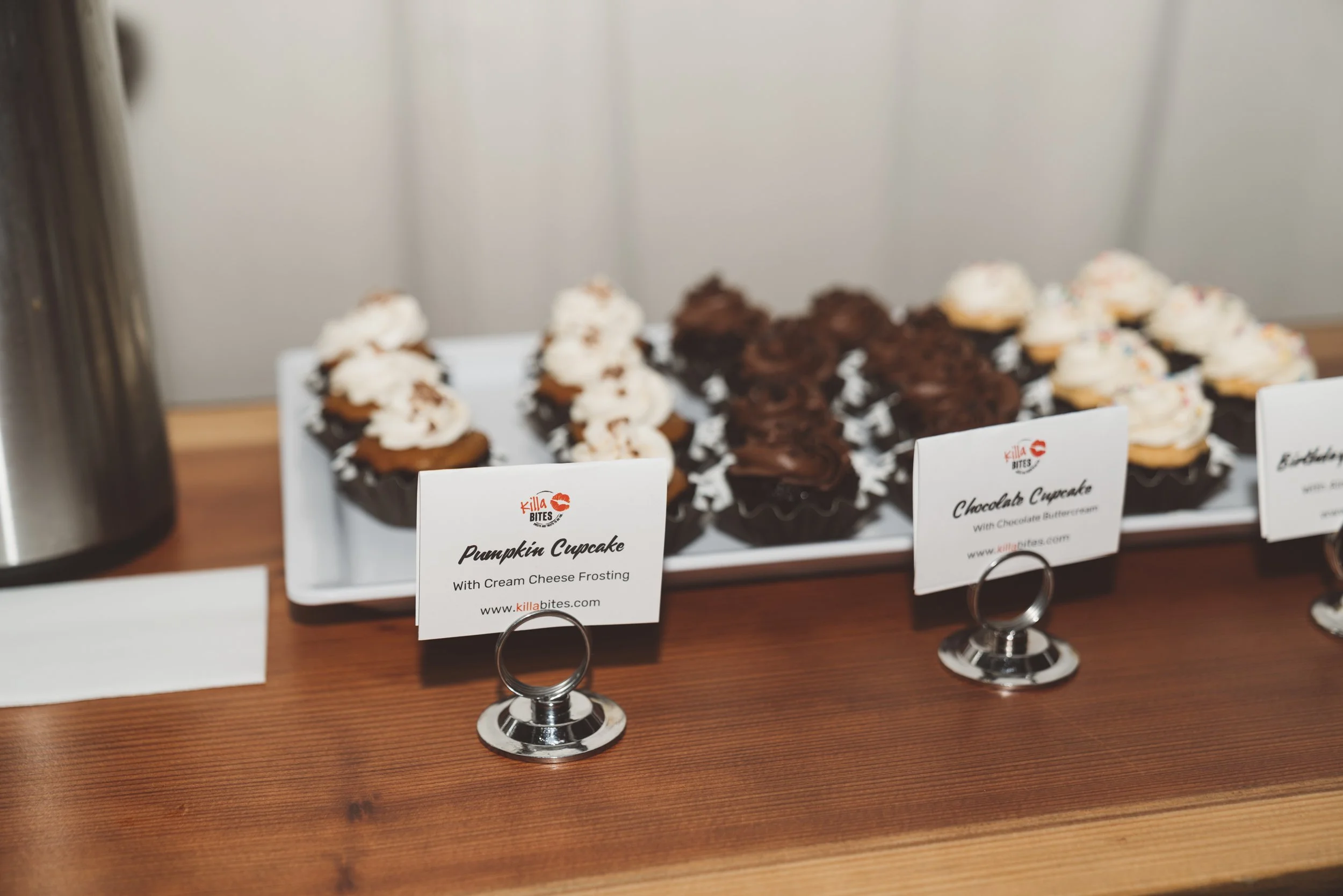 Assorted cupcakes displayed on a tray with a sign for pumpkin cupcakes with cream cheese frosting and chocolate cupcakes with chocolate buttercream.