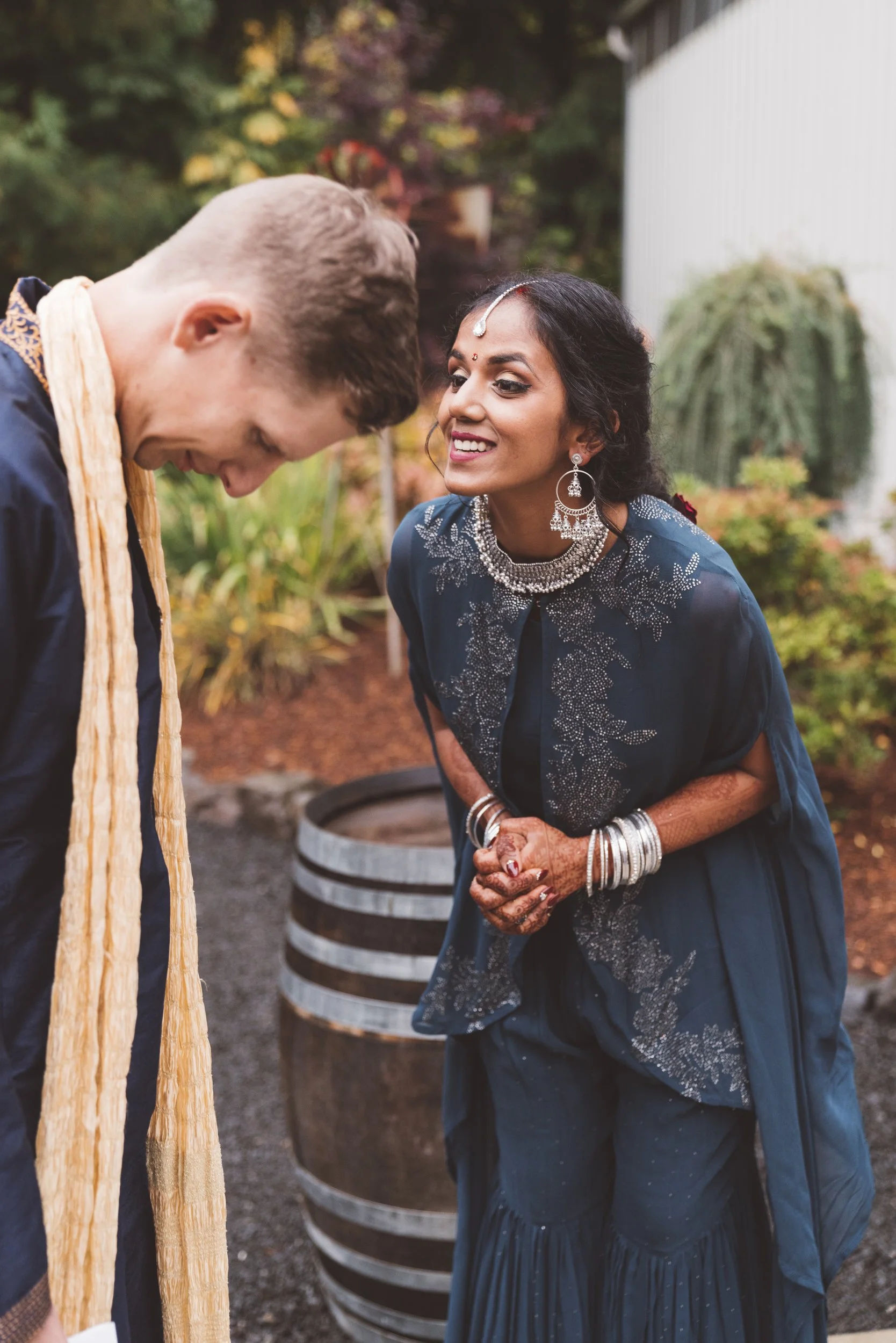 A woman dressed in traditional Indian attire with jewelry, leaning slightly forward and smiling at a man who is bowing his head, outdoors with greenery and a large barrel in the background.