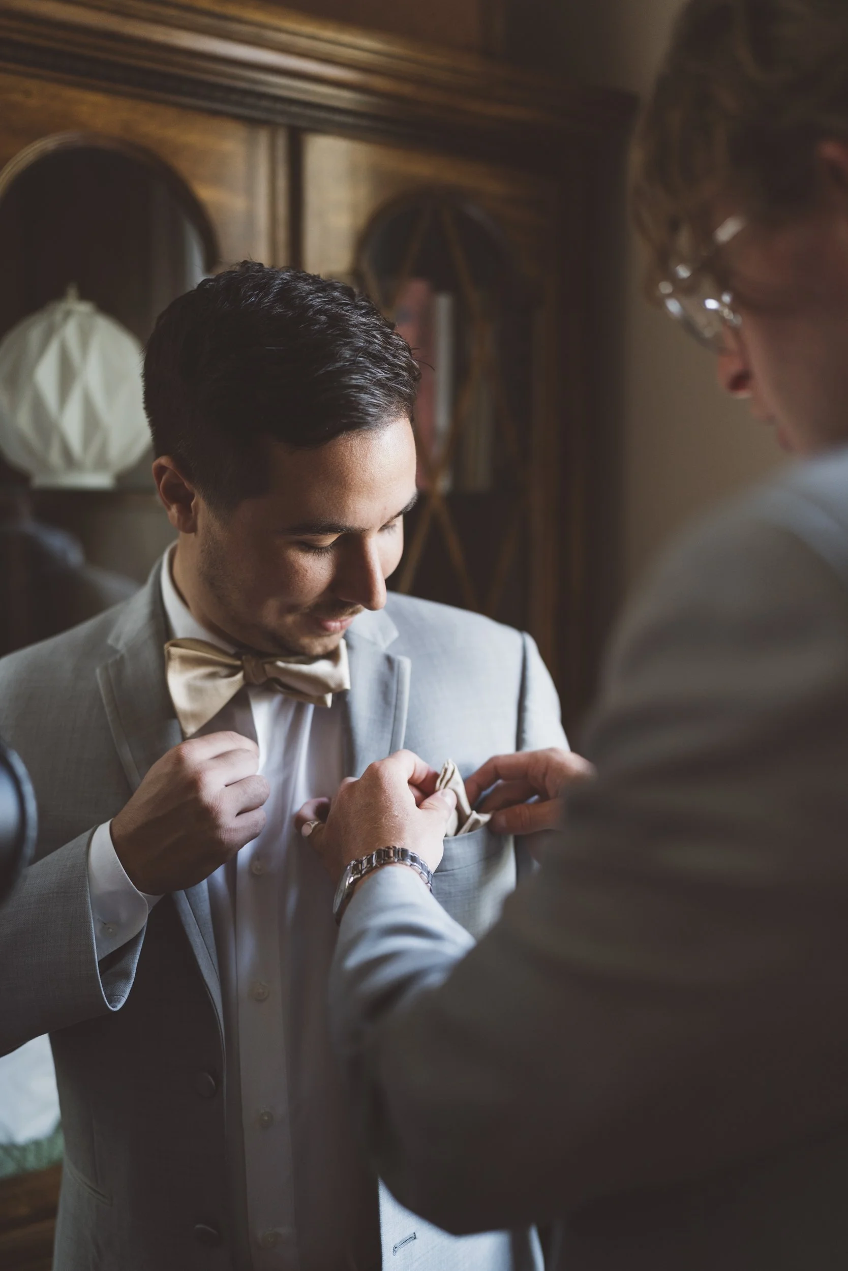 A groom in a light gray suit with a bow tie is being assisted with his pocket square by another man in a similar suit. The setting appears to be a room with wooden furniture and a mirror in the background.