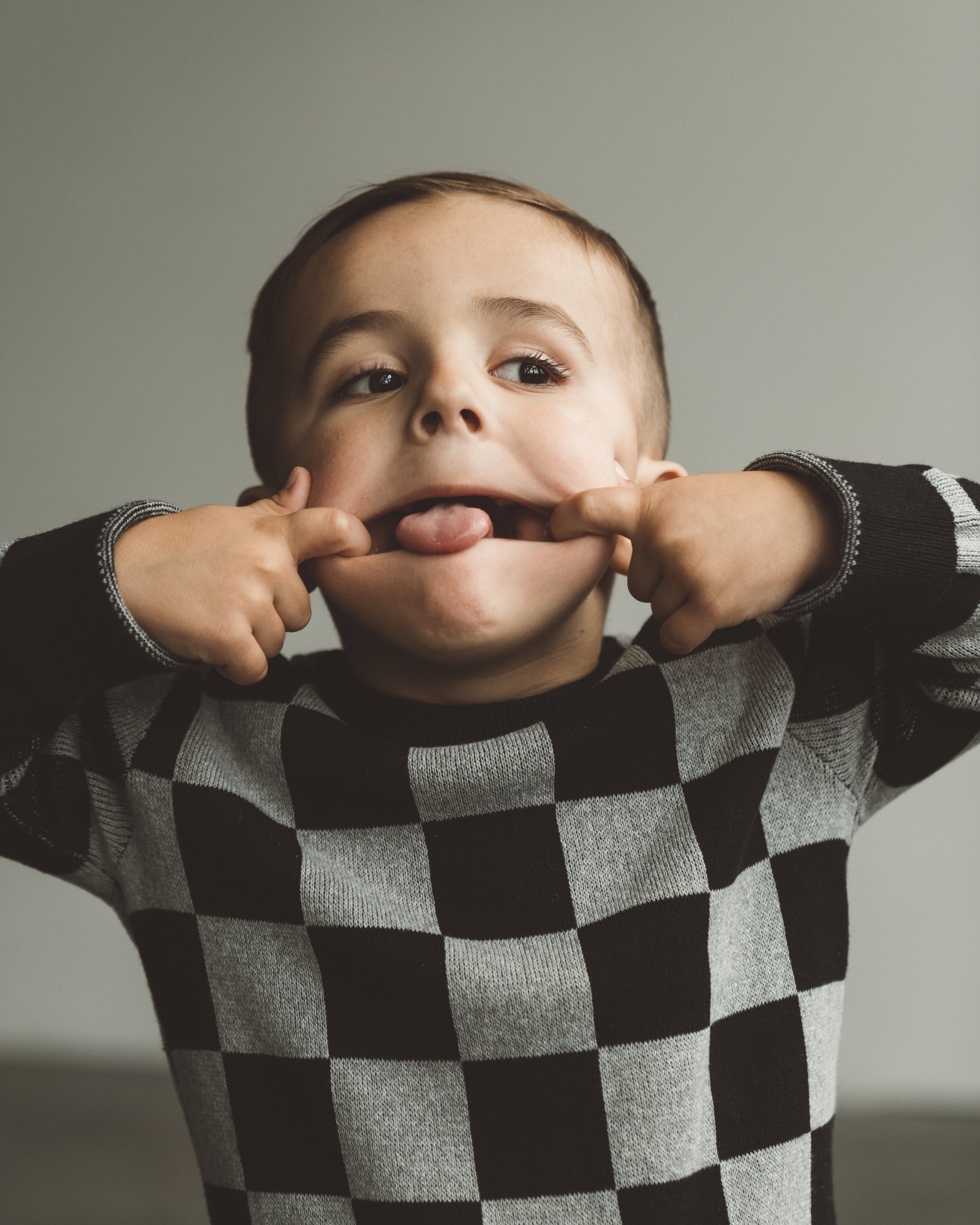 Child making a funny face, pulling cheeks, sticking out tongue, wearing a black and gray checkered sweater.