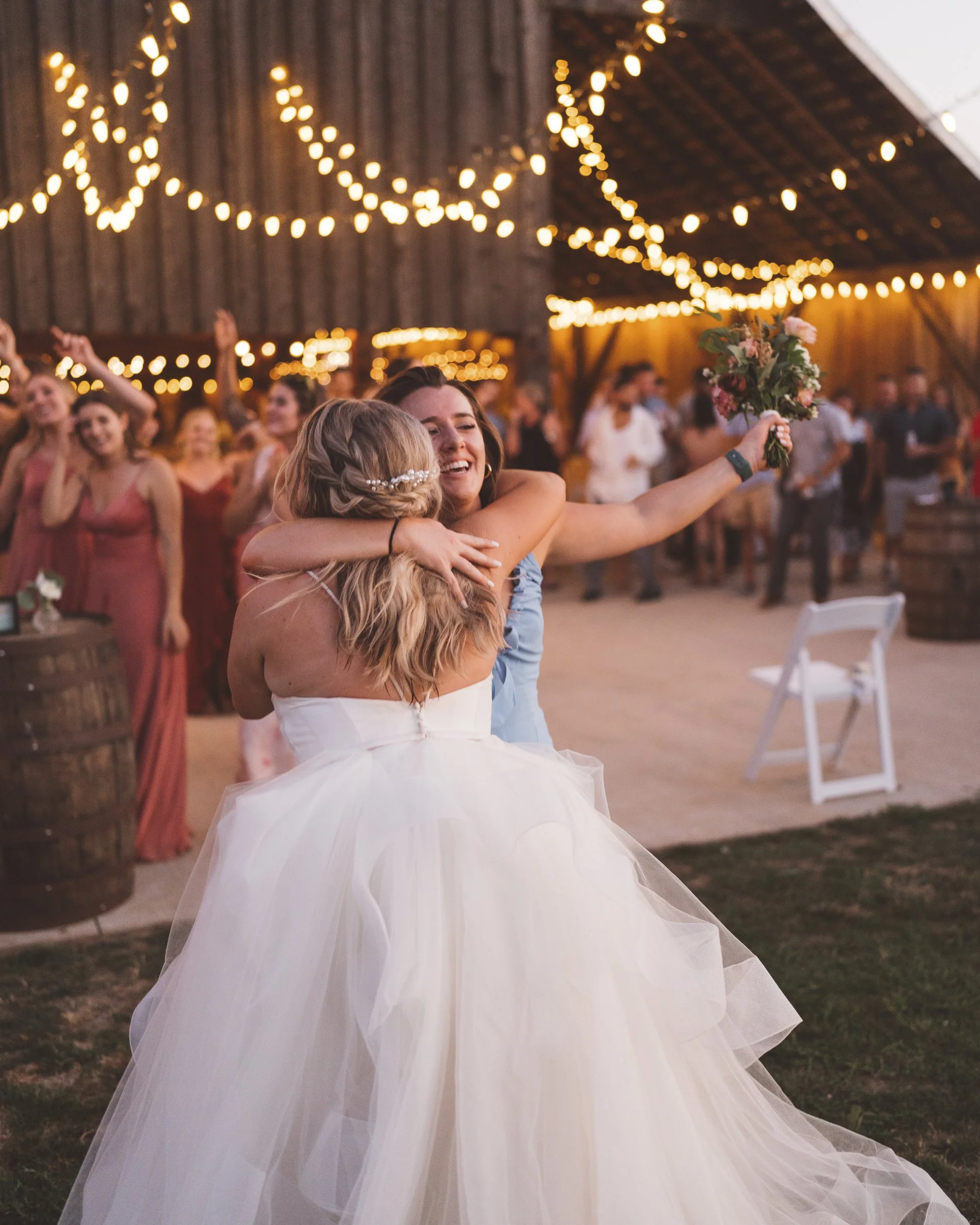 A joyful moment at a wedding reception with two women hugging, one in a wedding dress, the other in a blue dress, with string lights and dancing guests in the background.