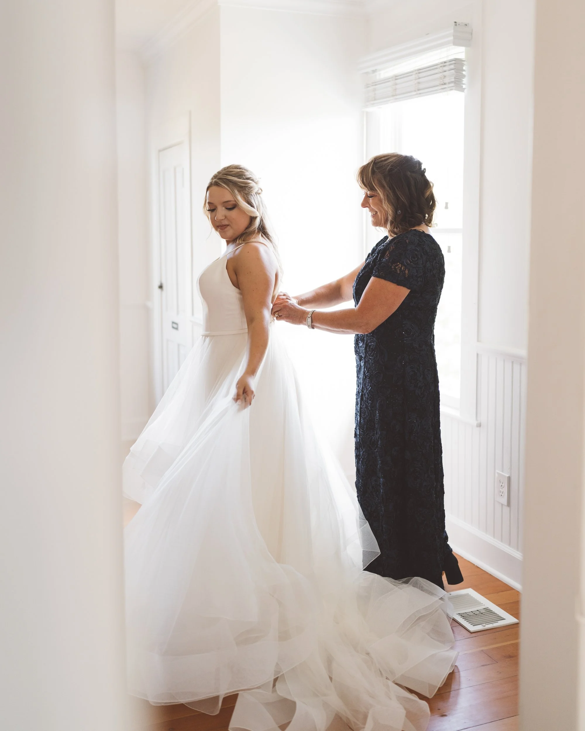 A bride in a white wedding gown is having her dress adjusted by a woman in a dark lace dress in a brightly lit room with white walls and a window with blinds.