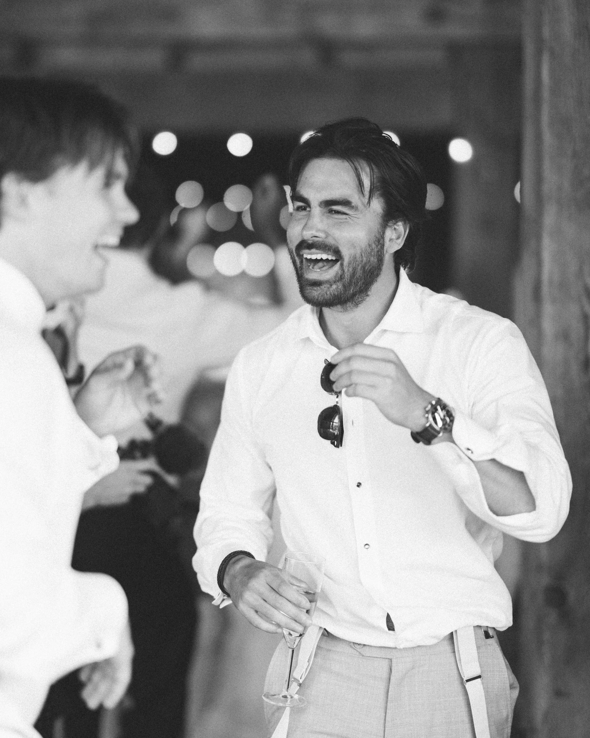 A man in a white shirt holding a glass of wine, laughing and talking to someone during a social gathering.