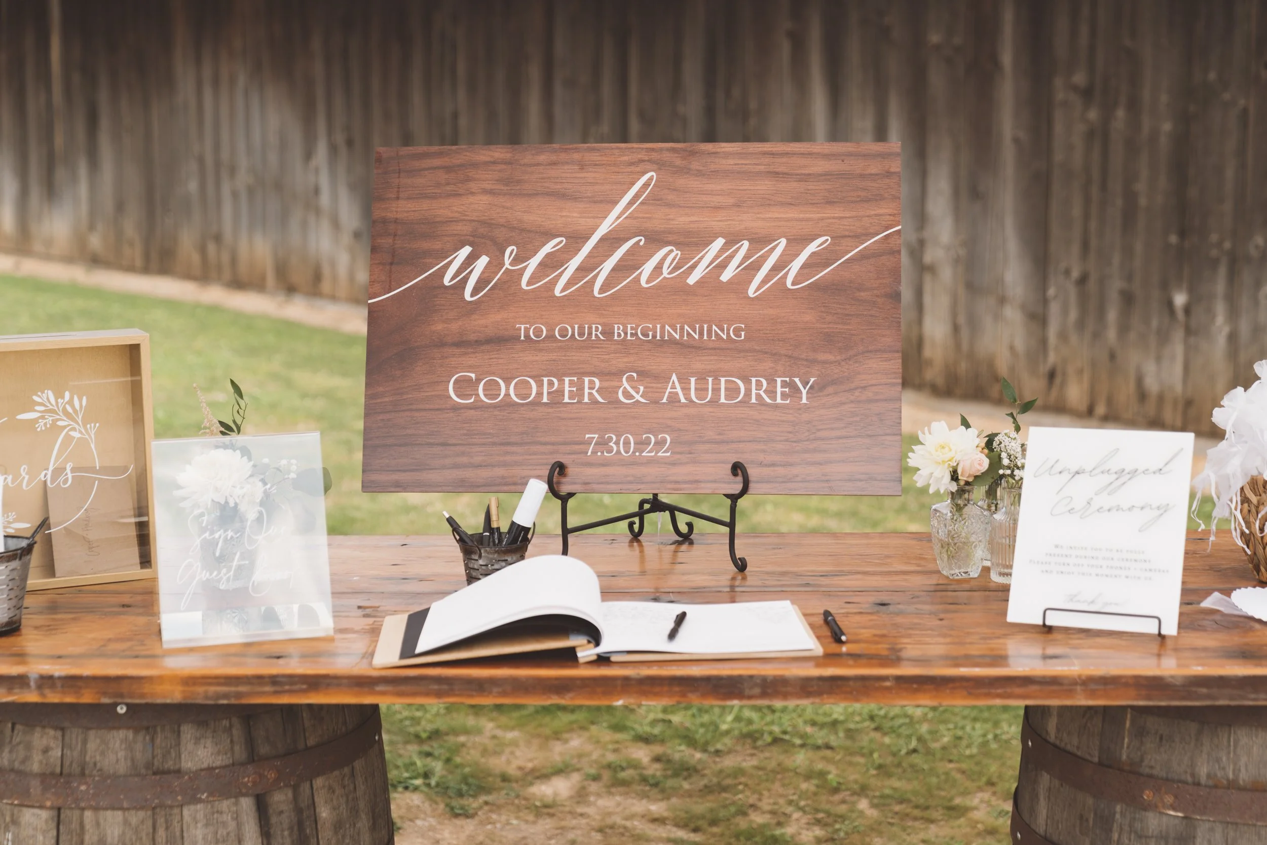 Wedding welcome sign for Cooper and Audrey with date 7.30.22 on a table with guest book, pens, and floral decorations, outdoors with a wooden fence in the background.