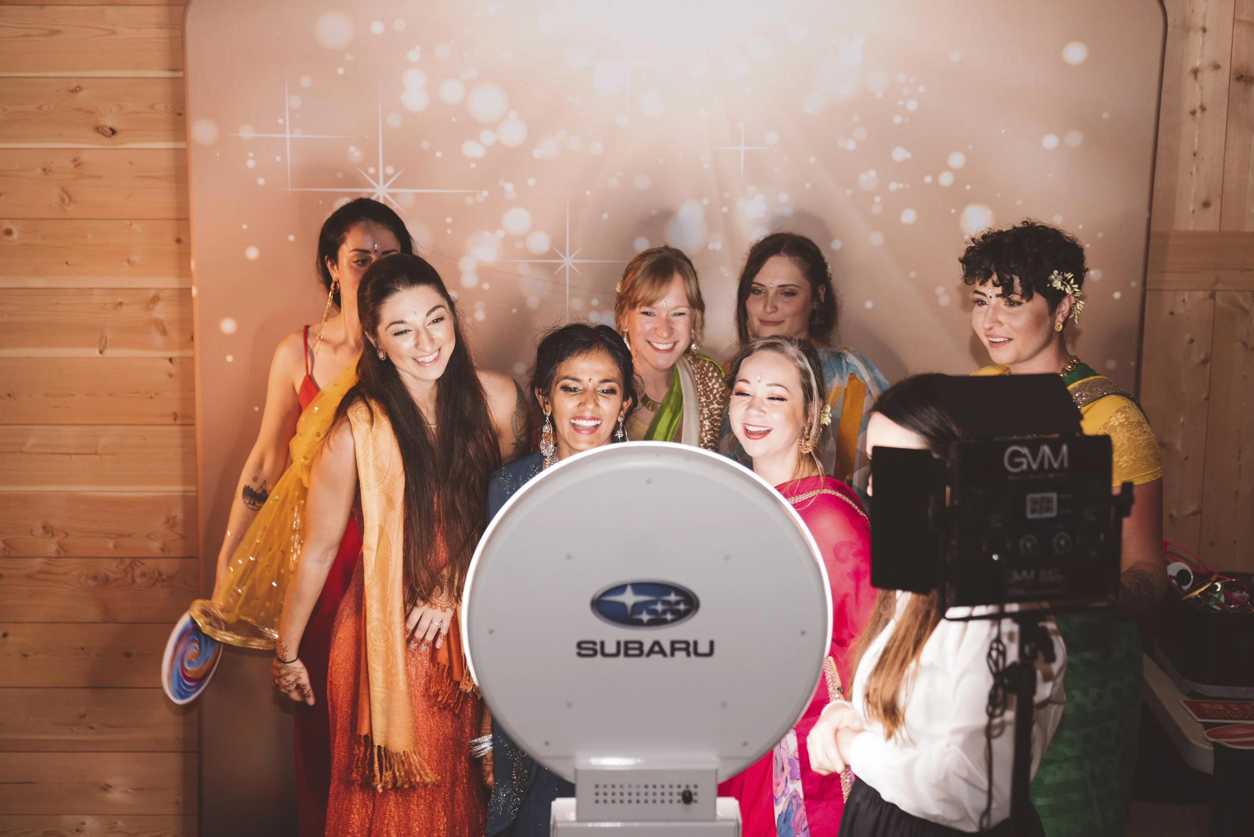 Group of women dressed in colorful traditional Indian attire taking a selfie in front of a camera with a Subaru logo, decorated with a wooden wall and starry backdrop.