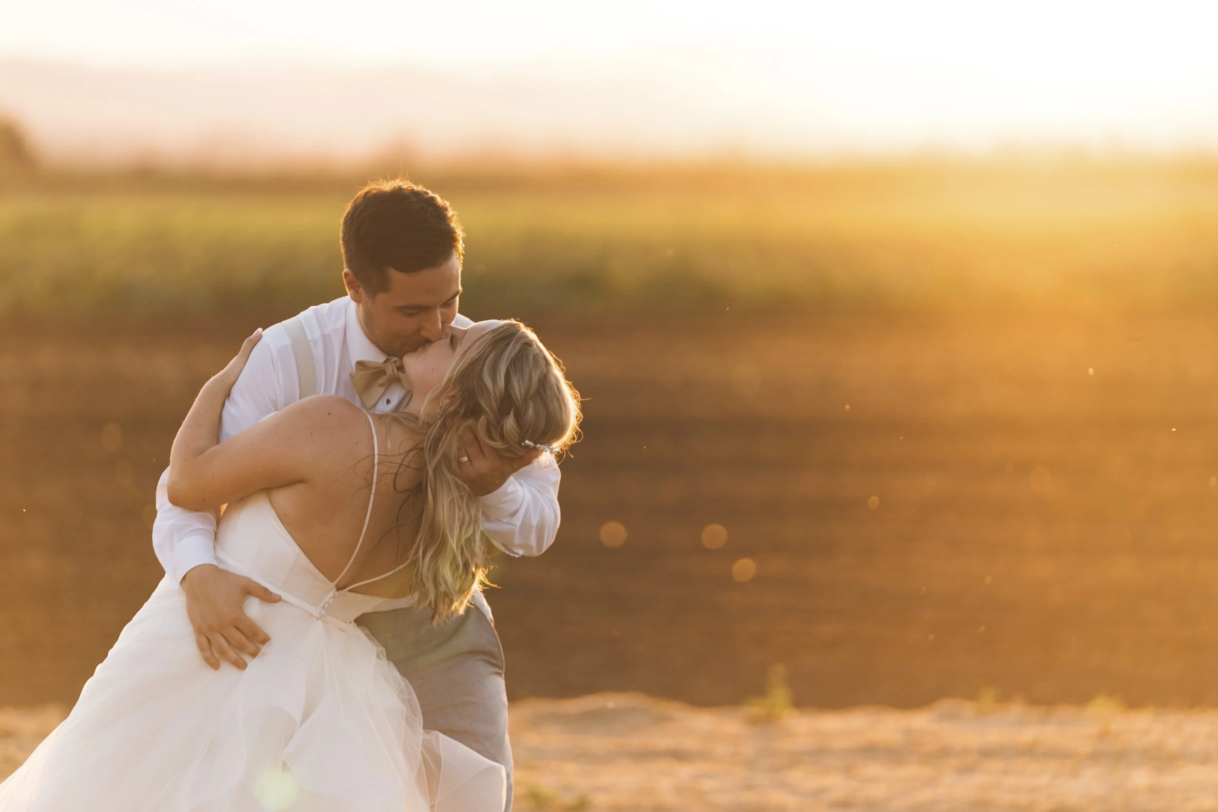 A newlywed couple sharing a kiss outdoors during sunset, with the groom dipping the bride as she leans back.