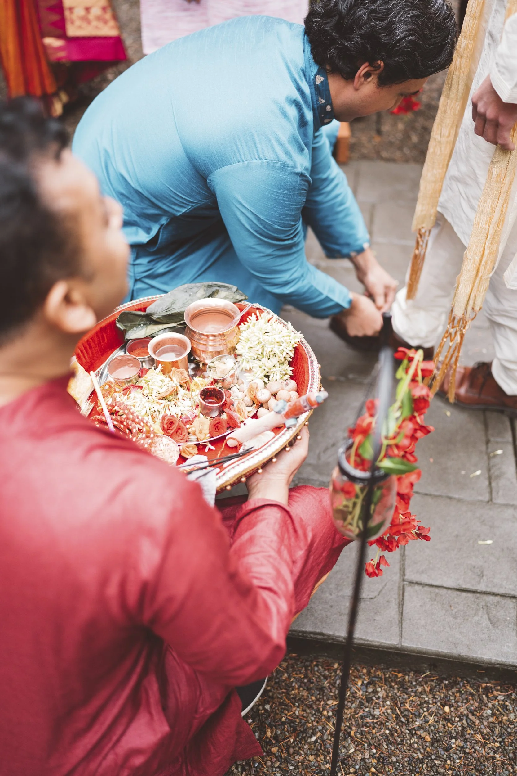 A traditional Hindu wedding ceremony with a priest holding a tray with offerings, flowers, and small cups, while a man is performing a ritual, tying a knot on a rope.