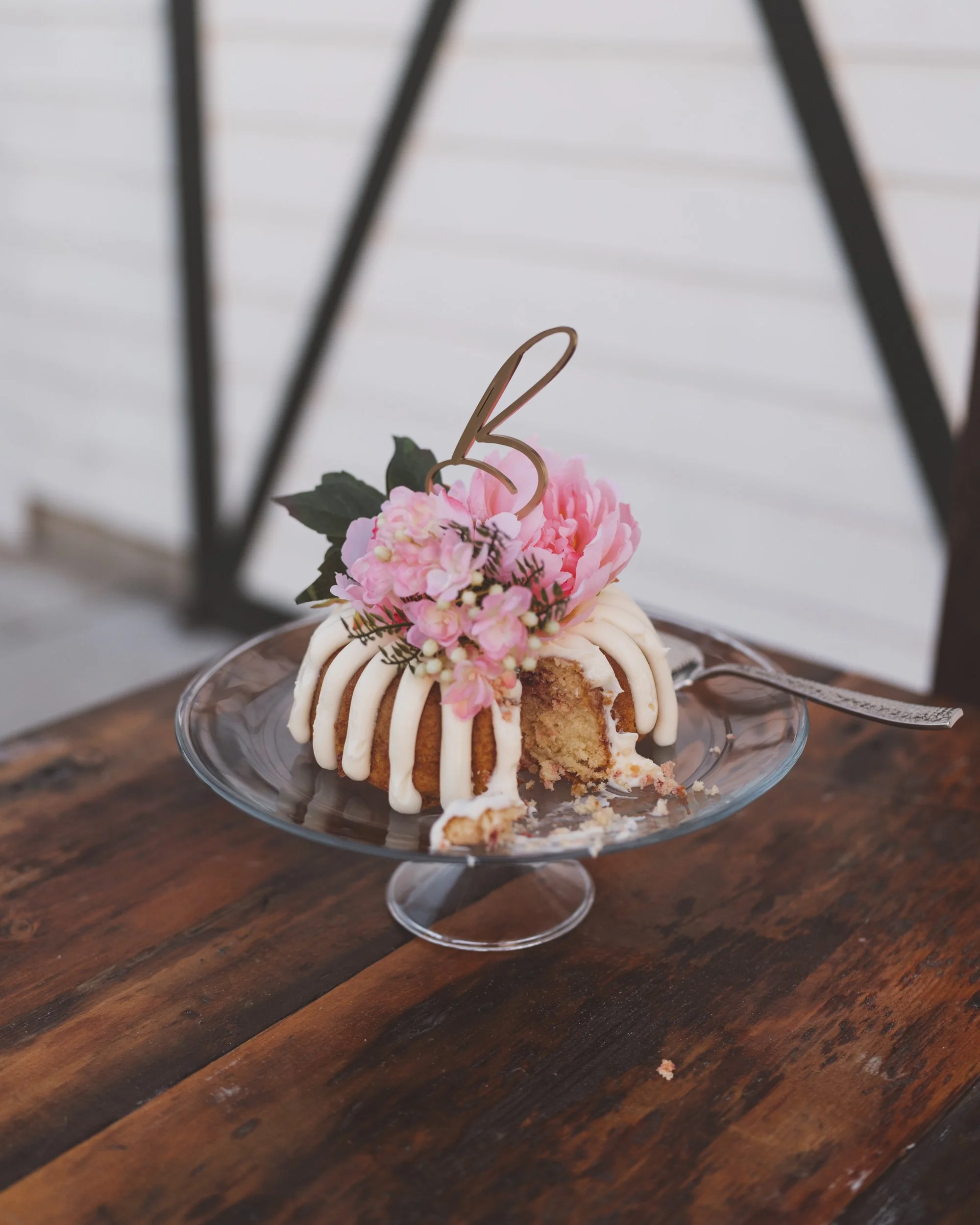 Close-up of a partially eaten birthday bundt cake decorated with pink flowers, greenery, and a gold '3' cake topper on a glass cake stand.