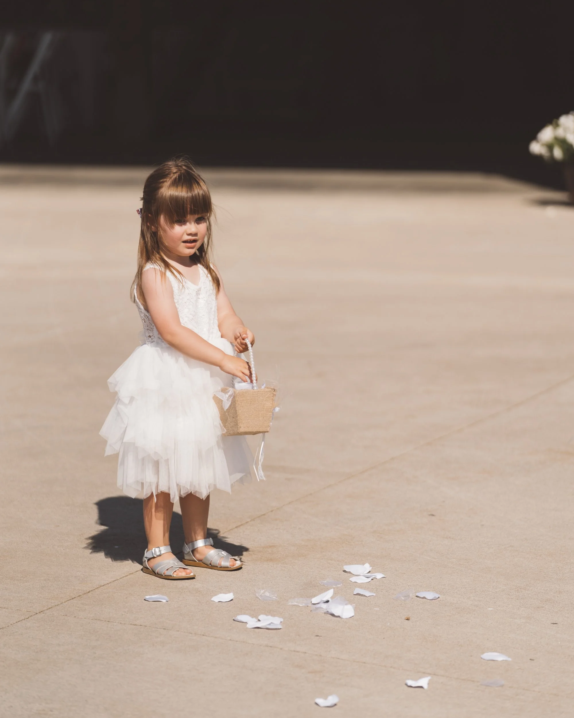 A young girl in a white dress holding a small basket with flower petals scattered on the ground nearby.