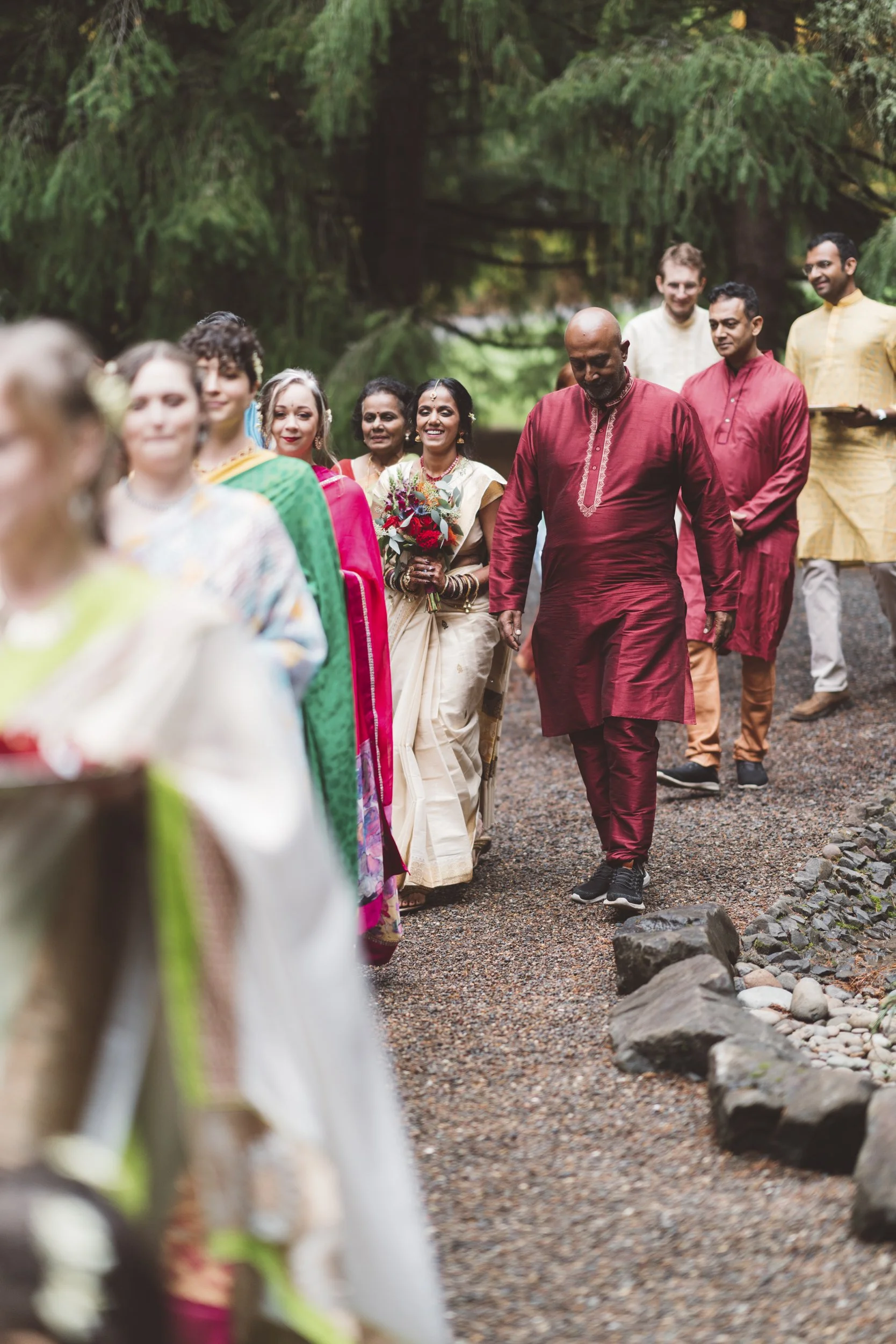 Group of people dressed in colorful traditional attire walking outdoors on a wooded path during a cultural or wedding celebration.