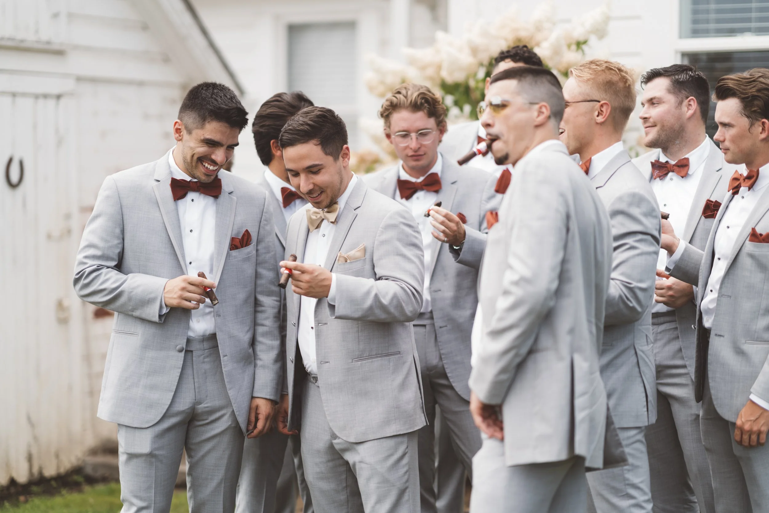 Group of men dressed in light gray suits with bow ties, standing outdoors on a porch, holding cigars, and engaging in conversation, at a wedding or formal event.