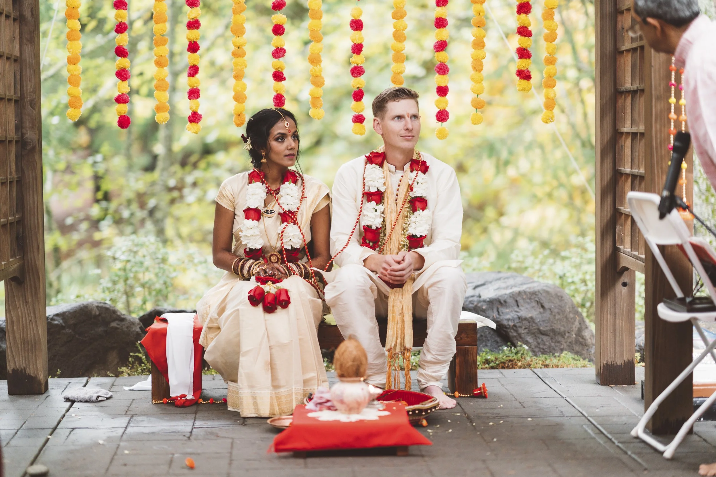 A couple participating in a traditional Indian wedding ceremony, seated outdoors under decorated hanging marigolds, wearing ceremonial attire and garlands, with a priest or officiant standing to the right.