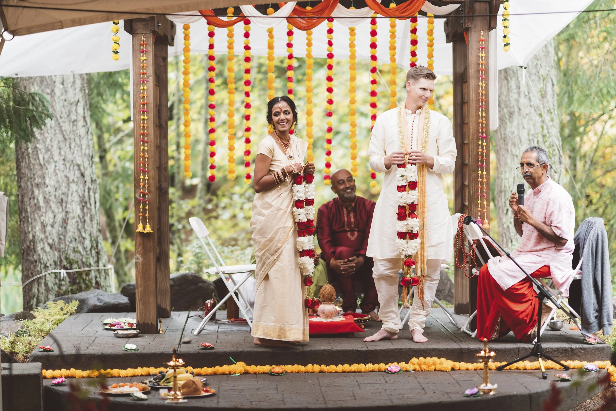 An outdoor Indian wedding ceremony with a decorated wooden canopy adorned with marigold and flower garlands. A bride and groom dressed in traditional Indian attire are holding flower garlands, standing on a platform decorated with marigold flowers. A