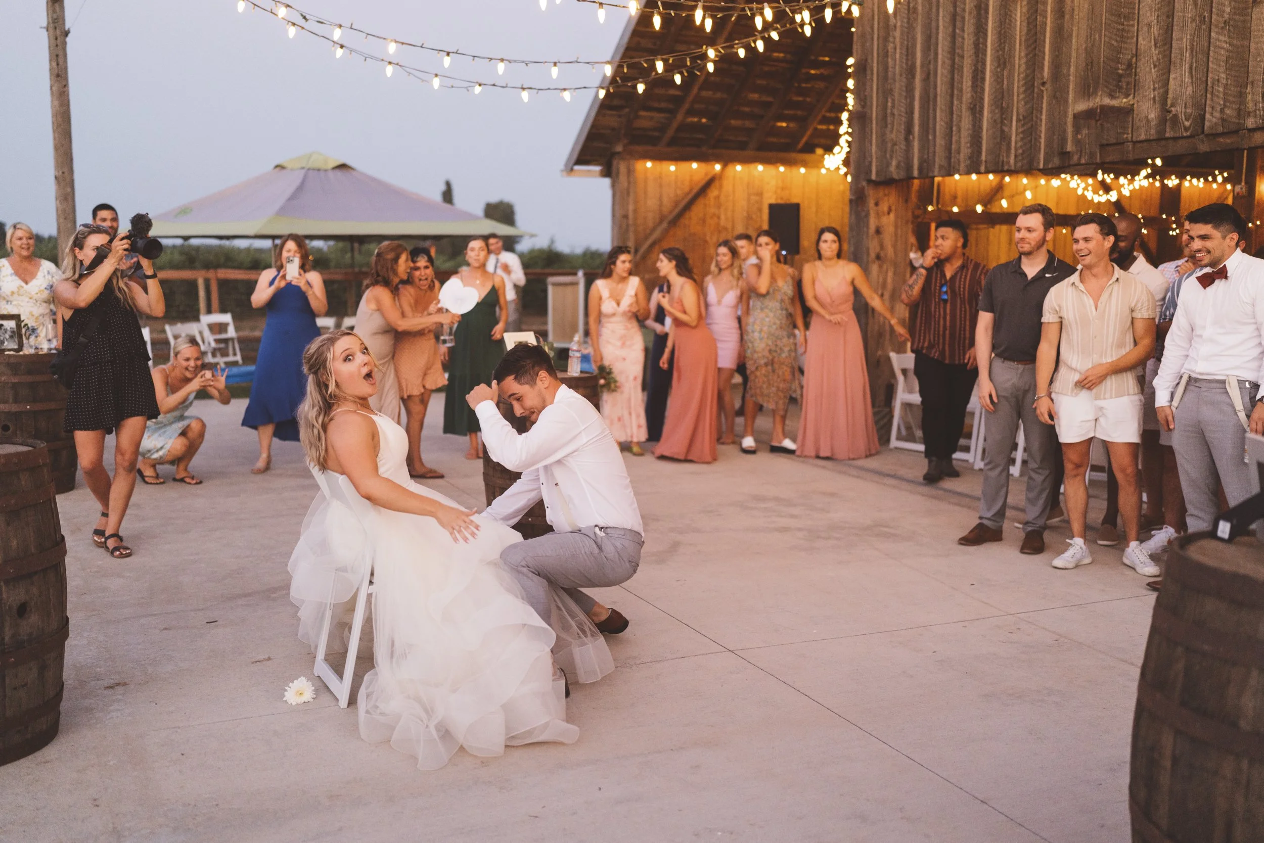 A wedding reception with the bride and groom dancing in the center, surrounded by guests. The bride is wearing a white dress, and the groom is in a white shirt and gray pants. Guests are watching, taking photos, and standing near string lights and wo