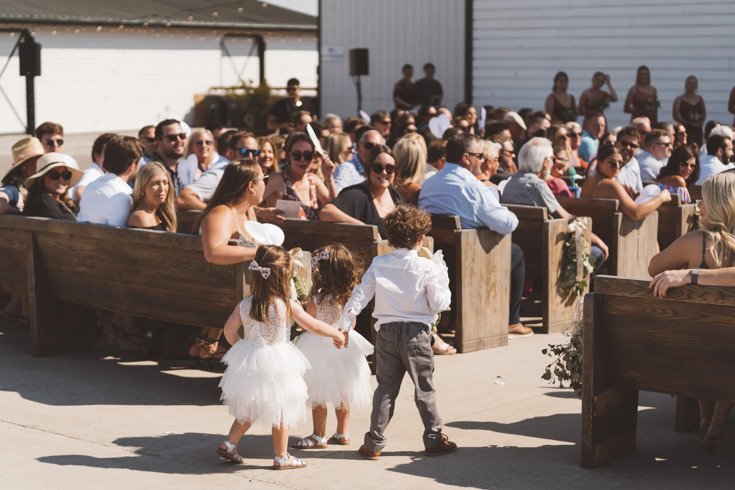 Children in white dresses and a boy in gray pants walk hand-in-hand in front of seated guests at an outdoor wedding ceremony on a sunny day.