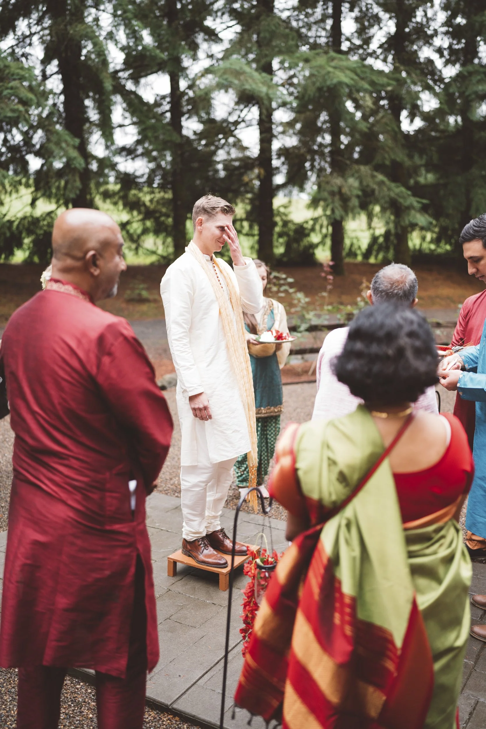 A man in traditional Indian attire standing on a small platform during a ceremony surrounded by people, in an outdoor setting with trees in the background.