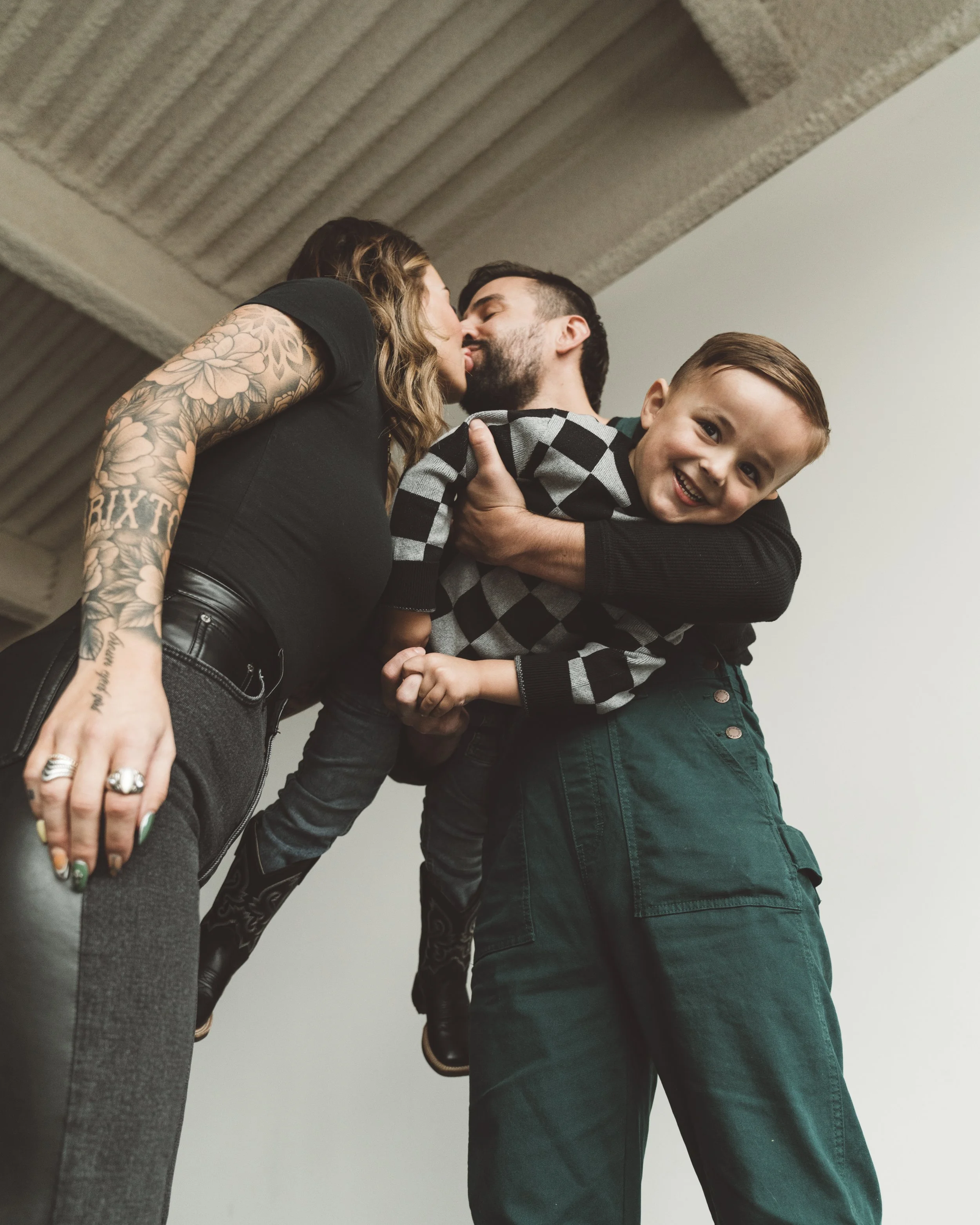 A young family of three sharing a happy moment indoors, with the mother and father kissing and their young son smiling, being held by the father.