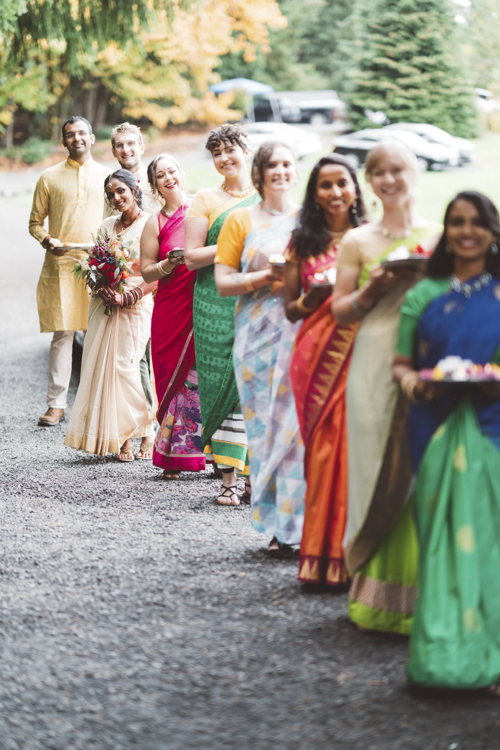 A diverse group of people in traditional Indian attire standing in a line outdoors, holding plates with food and flowers, with colorful trees and parked cars in the background.