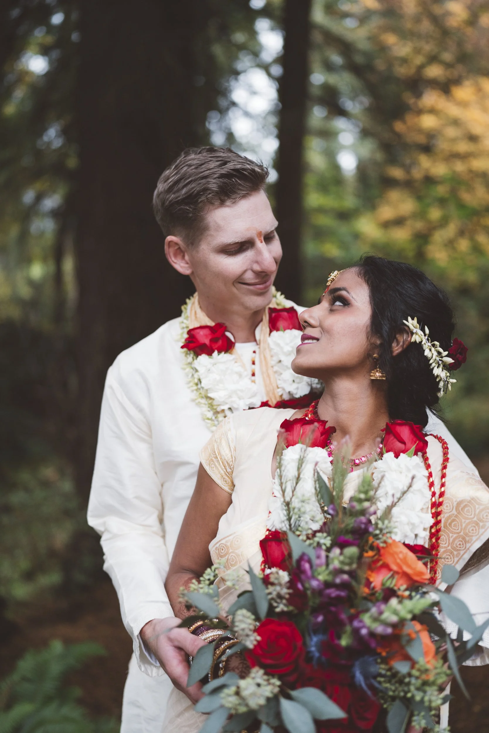 A couple in traditional Indian attire shares a moment in a forest, with the woman holding a large bouquet of colorful flowers.
