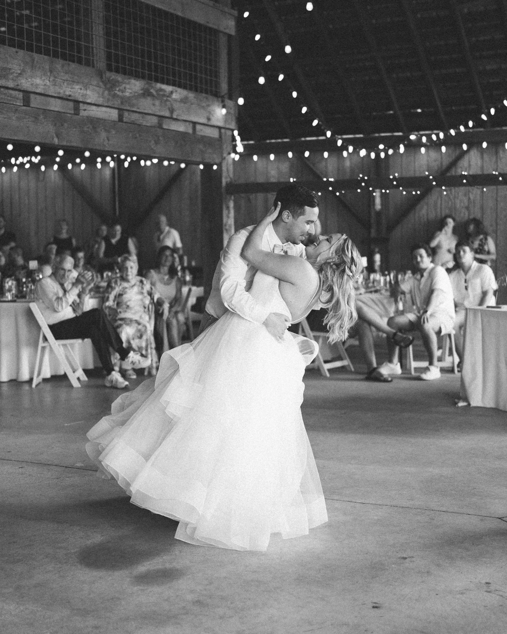 A bride and groom sharing a dance at their wedding reception, with guests seated at tables in the background, in a rustic barn decorated with string lights.
