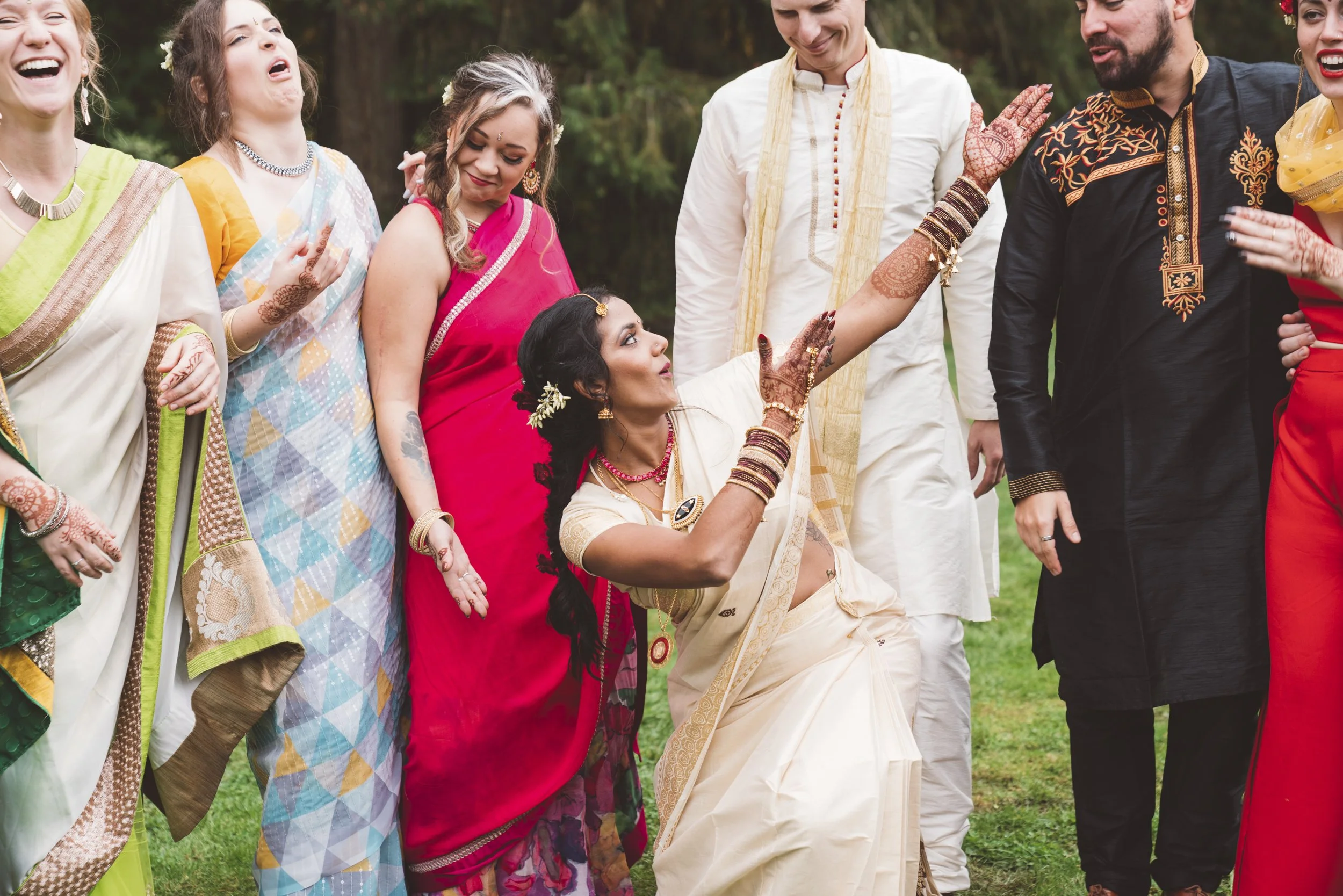 A group of people dressed in traditional Indian clothing, celebrating outdoors. The woman in a cream sari is performing a dance or ritual, surrounded by others who are smiling and watching.